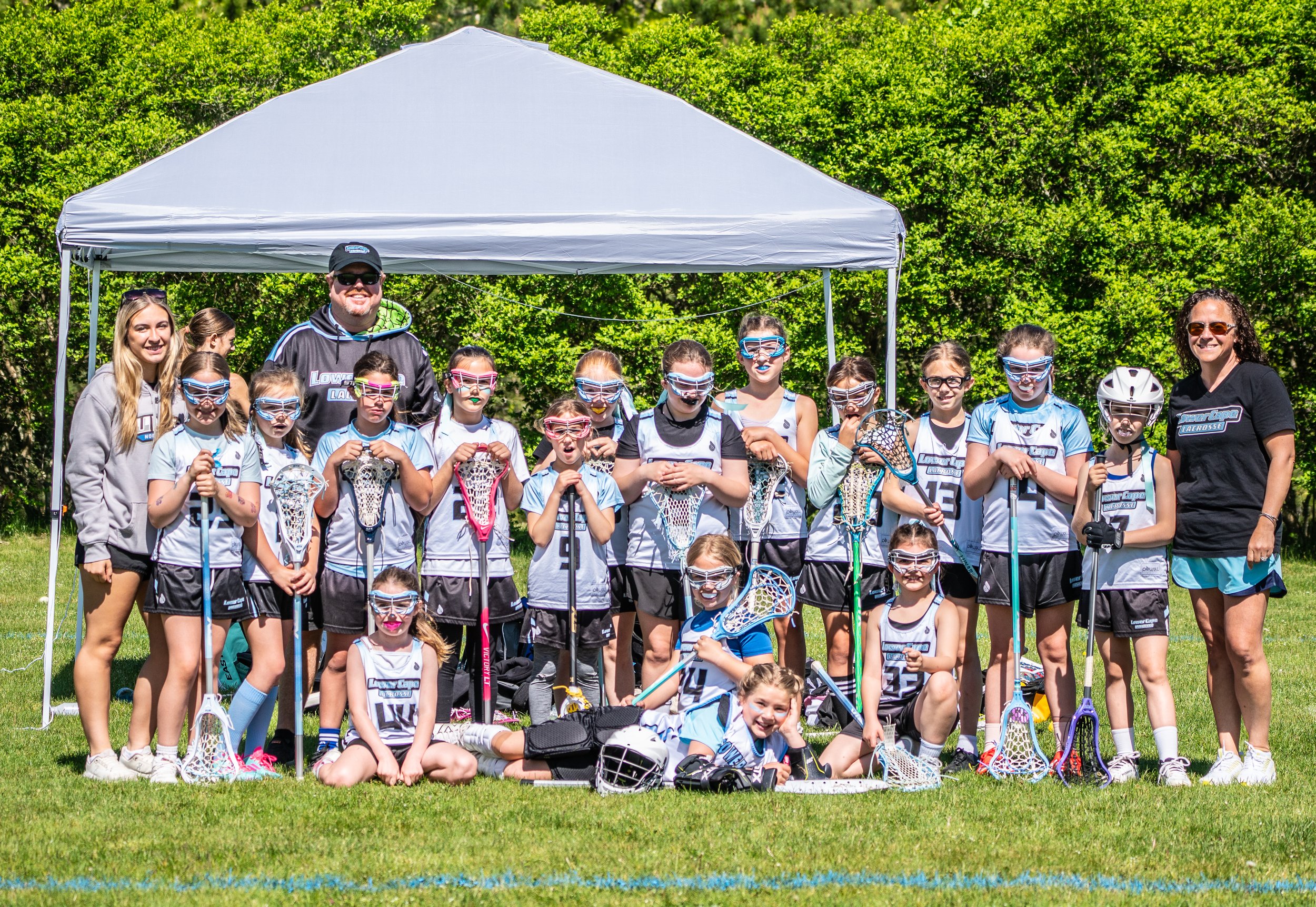 Group photo of a girls' youth lacrosse team outdoors near a grassy field with a white canopy for shade, led by two adult women coaches, all wearing team uniforms and protective goggles, holding lacrosse sticks, some sitting or lying on the grass, with green trees in the background on a sunny day.