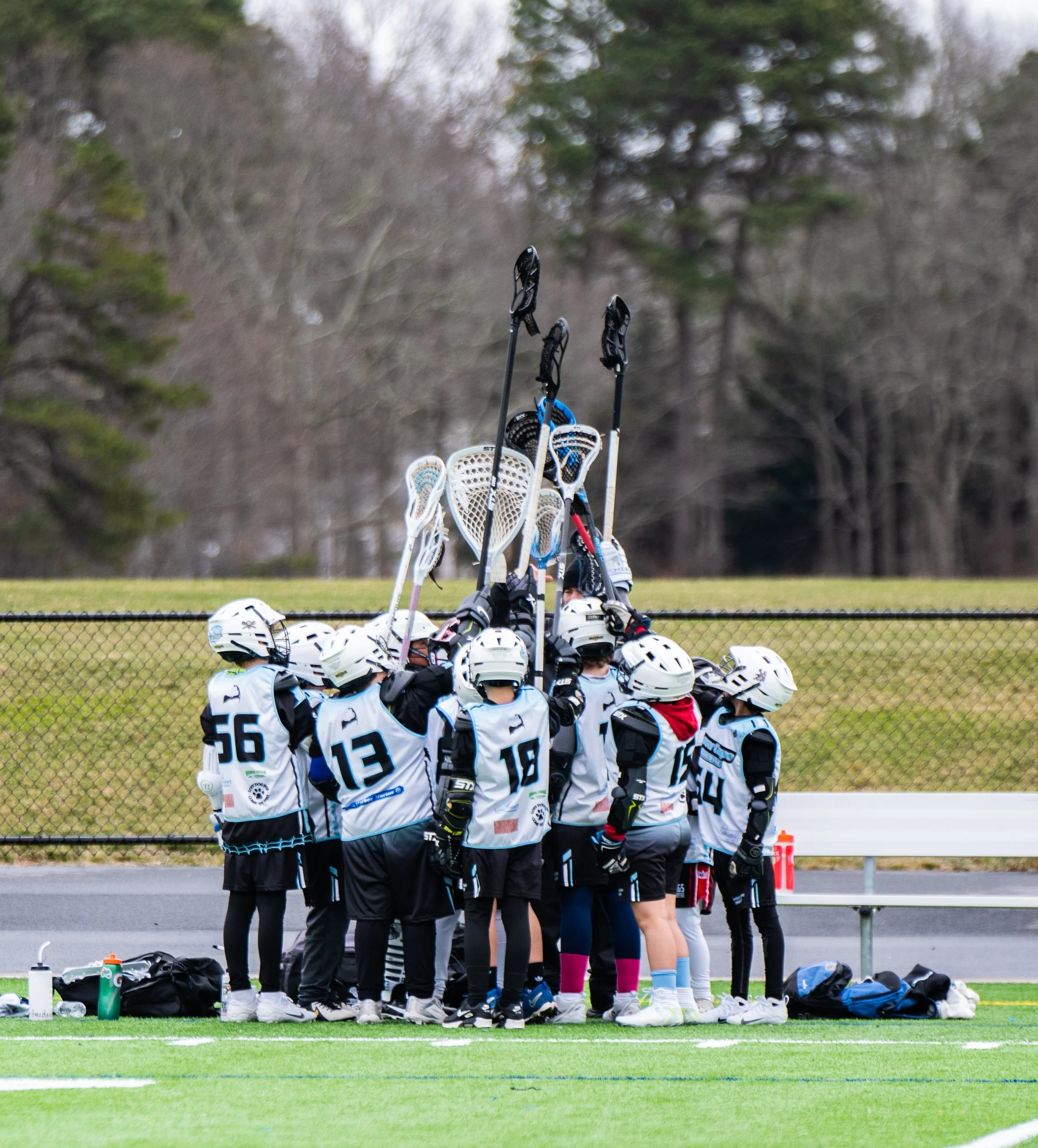Youth lacrosse team huddled together on the field with their sticks raised in a line, wearing helmets and uniforms, during a game.