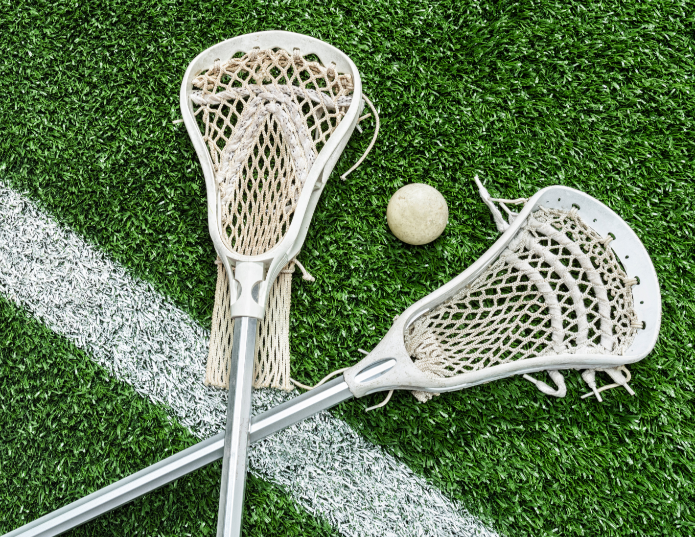 Two lacrosse sticks crossed on a green turf field with a white line and a lacrosse ball nearby.