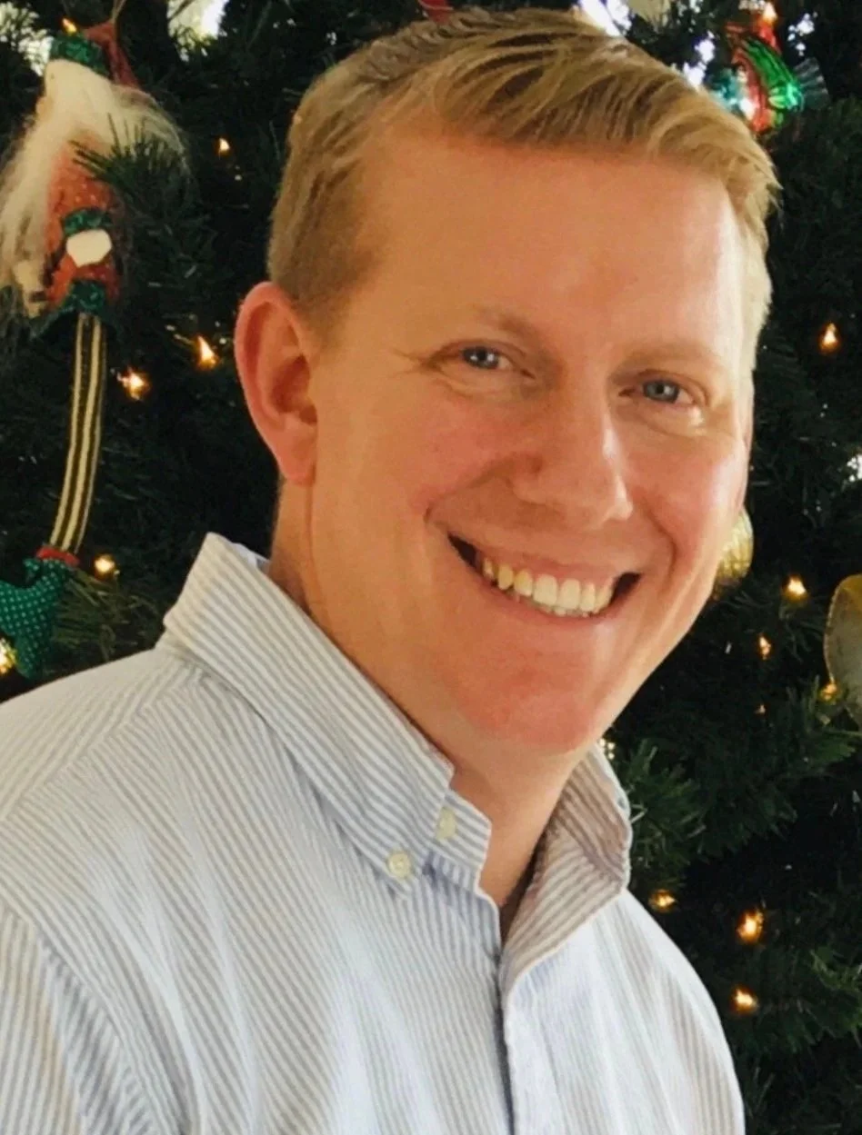 A smiling man with blonde hair in front of a decorated Christmas tree.