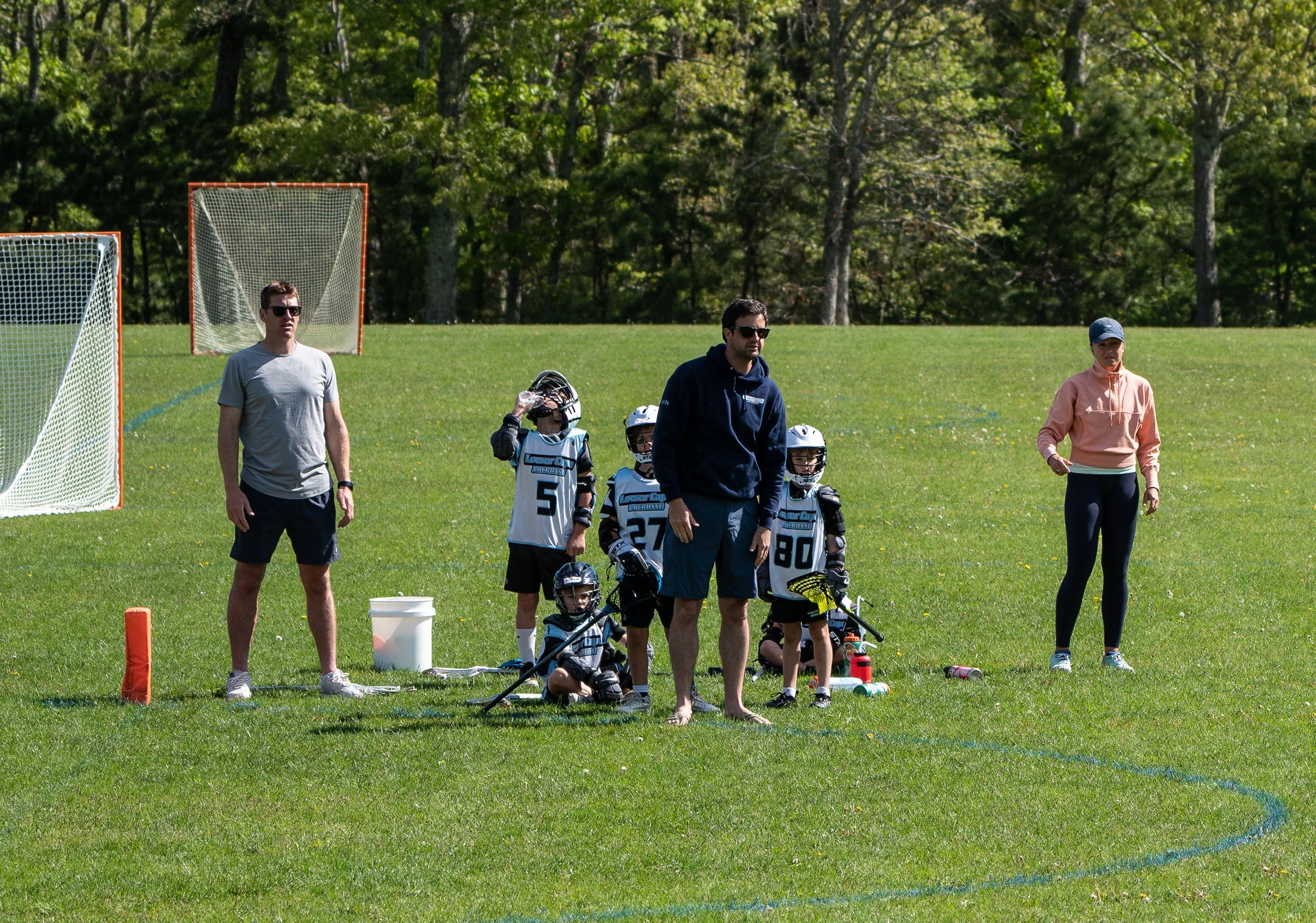 Youth lacrosse team wearing helmets and jerseys, with coaches on a grassy field during practice, surrounded by soccer goals and trees.