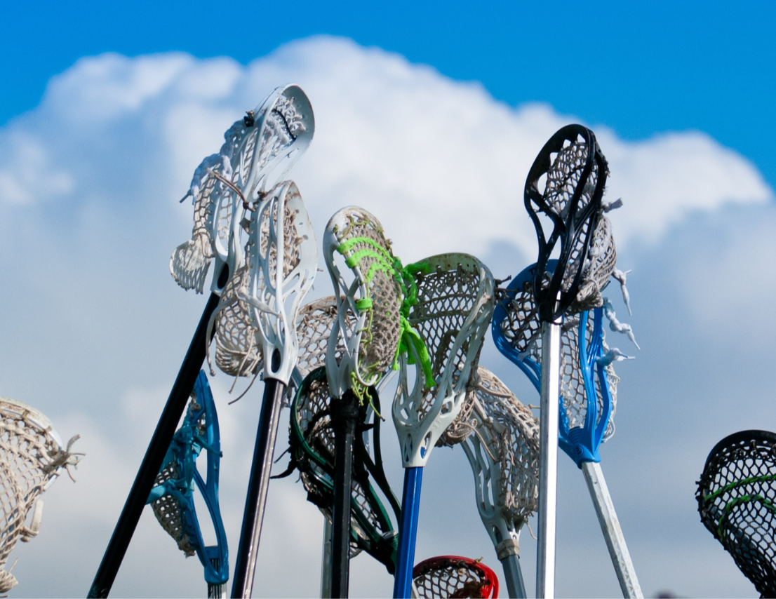 A group of lacrosse sticks with different colored heads is held upright against a background of a cloudy blue sky.