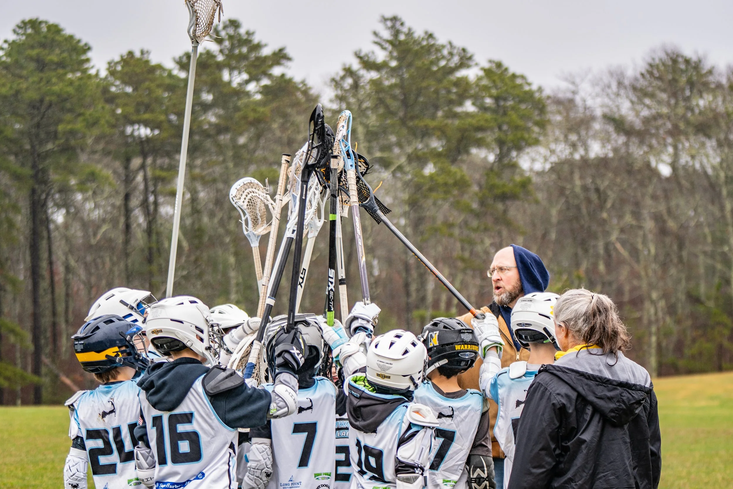 A group of high school boys lacrosse players wearing white helmets and jerseys gathered around two coaches in an outdoor field setting with trees in the background. The players are holding lacrosse sticks up towards the coaches.
