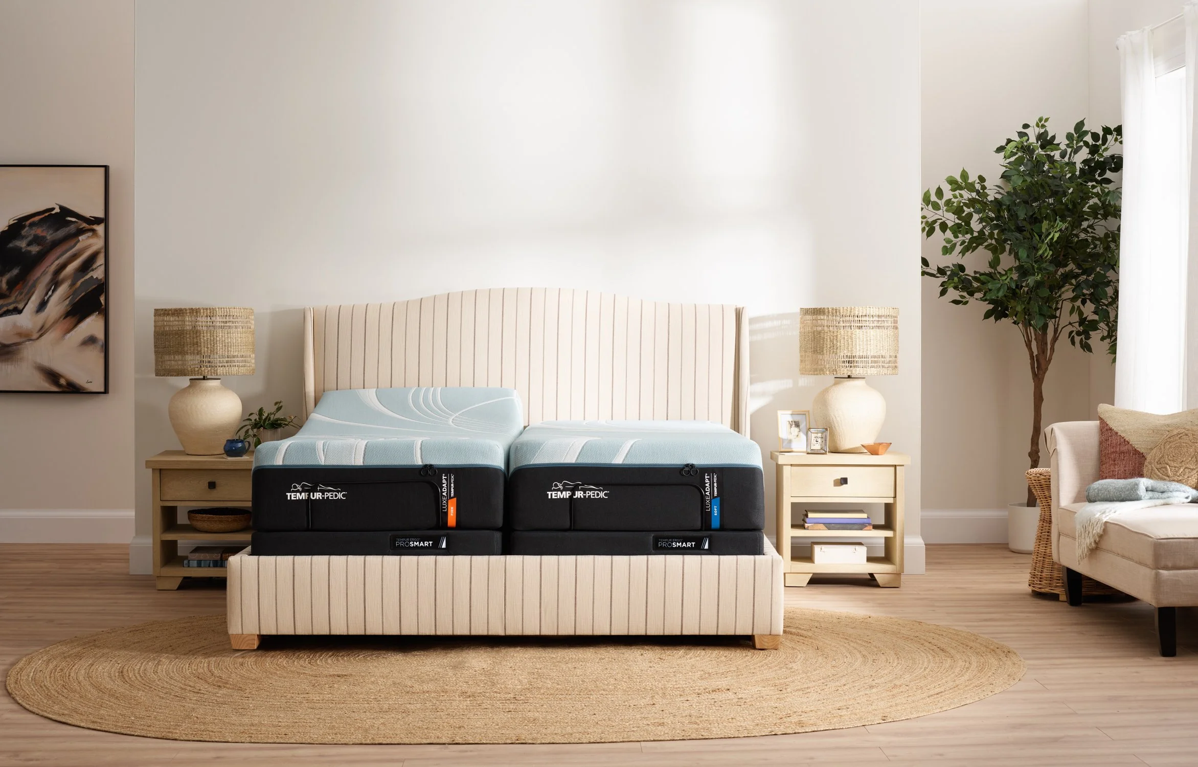 A bedroom with a large headboard against a plain wall, two nightstands with lamps, a framed abstract art piece on the wall, a potted plant, and a beige armchair. A mattress with Tempur-Pedic branding is placed on the bed frame in the room.