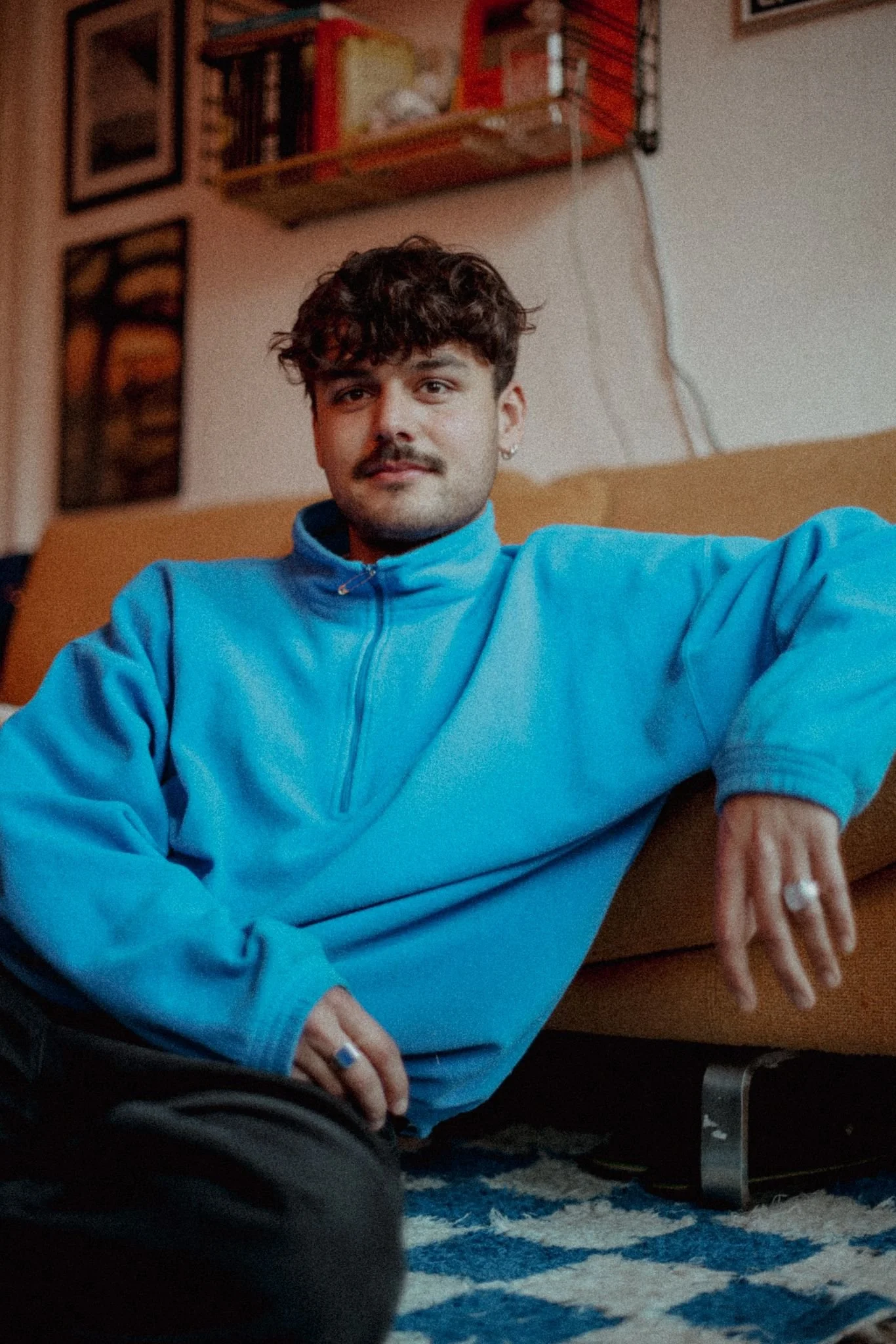A young man with curly hair and a mustache wearing a blue fleece quarter-zip pullover, sitting on a patterned blue and white rug. He is leaning against a beige sofa and has jewelry on his fingers, with framed pictures and a bookshelf on the wall behind him.