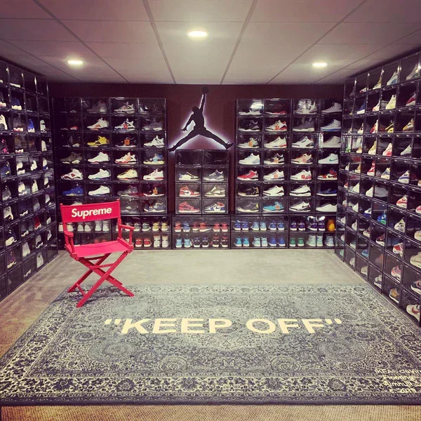 A shoe sneaker collection in a room with shelves filled with sneakers. A sale chair with a red Supreme logo is in front, and a rug on the floor reads 'KEEP OFF!'