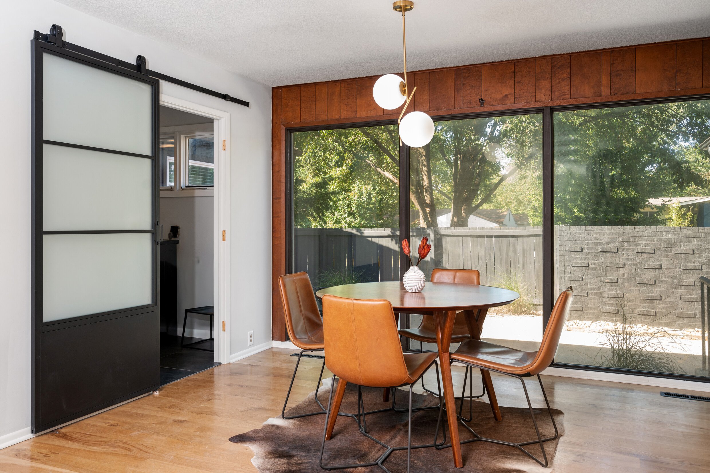 A dining room with a round wooden table and four brown leather chairs, a vase with red flowers on the table, a large window with greenery outside, a modern light fixture hanging above the table, and a sliding frosted glass door.