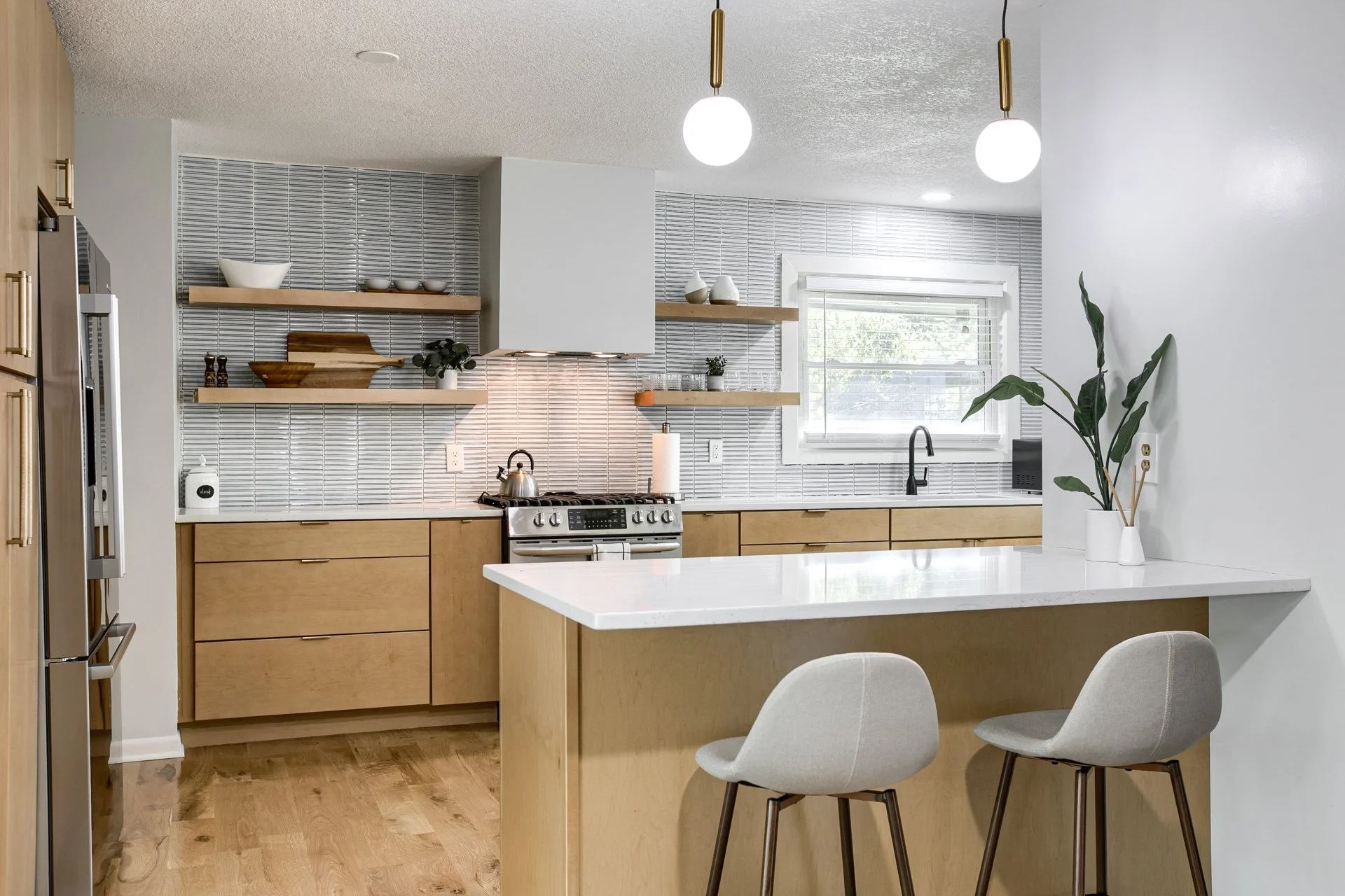Modern kitchen with white countertops, light wood cabinets, open wooden shelves with bowls and decorative items, stainless steel stove with kettle, window above sink, two white barstools, two globe pendant lights, potted plant on counter.