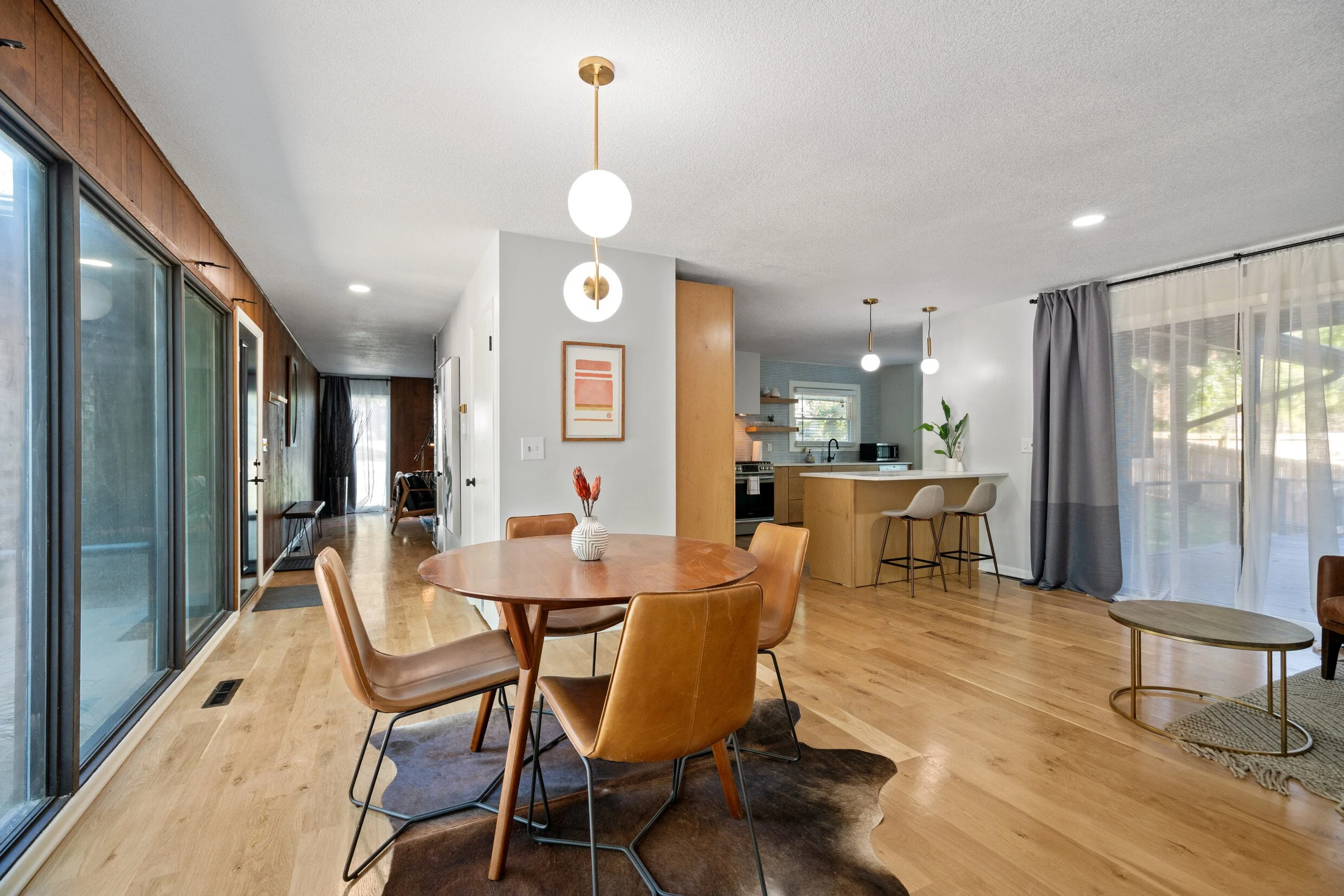 Open-concept living and dining area with wooden floors, a round dining table with five leather chairs, large glass sliding doors, a kitchen with a breakfast bar, and a sitting area in the background.