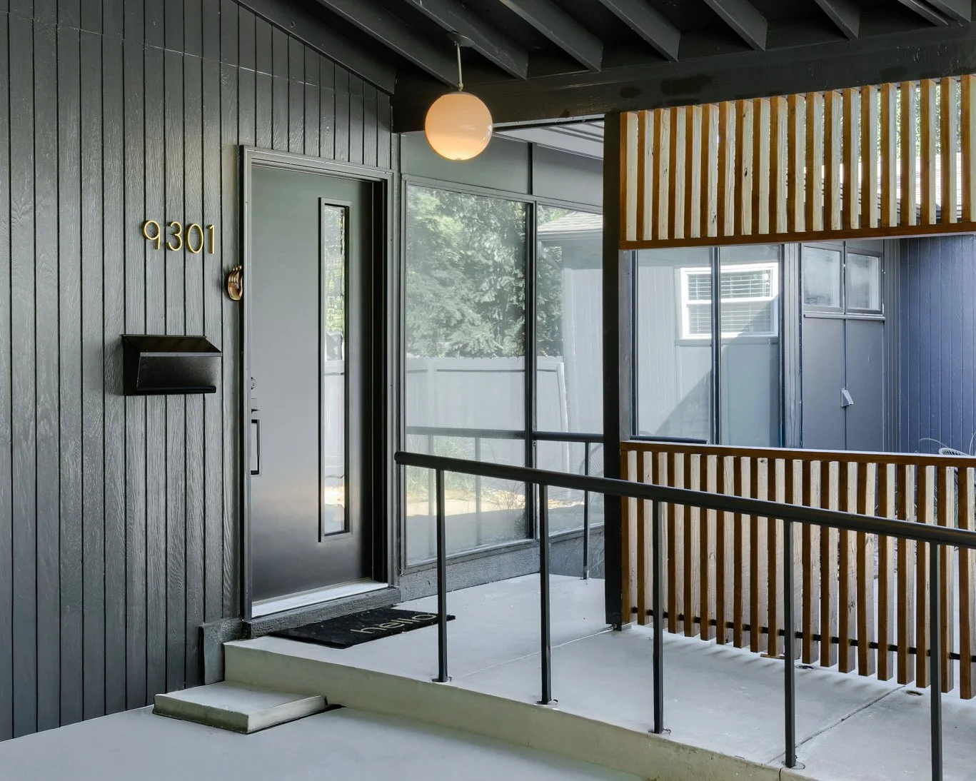 Apartment entrance with black door numbered 9301, black mailbox, black welcome mat, and a small step leading to the door. The area features dark vertical siding, a black ceiling, a round hanging light, and a wooden railing next to a large window.