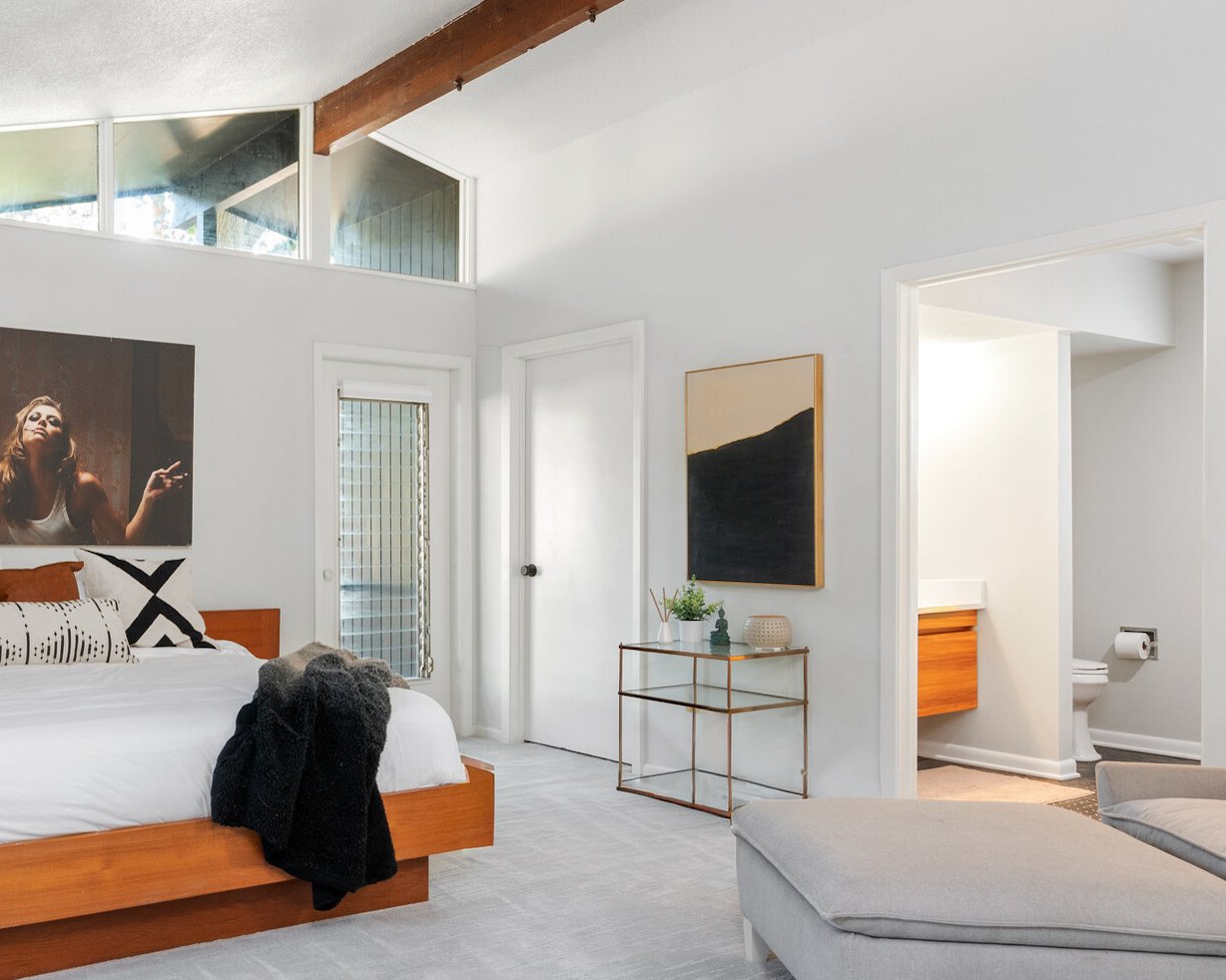 A modern bedroom with white walls, a wooden bed frame, and a black-and-white photograph above the bed. There is a door with a vertical window, a small gold metal table with decor, and a doorway leading to a bathroom. The room features a sloped ceilin