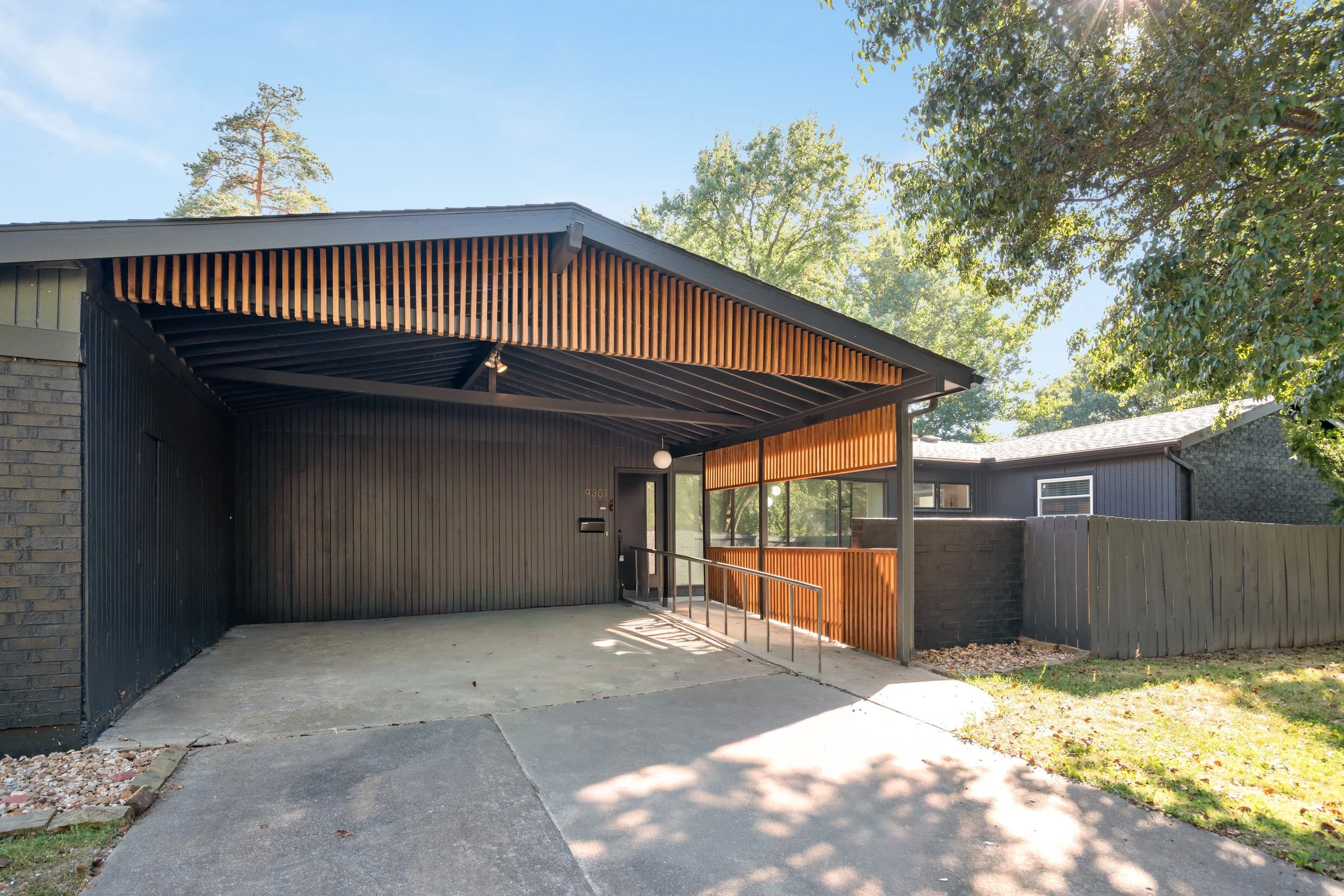 Modern house with black exterior, attached carport with a wood slat design, concrete driveway, and a small grassy yard with trees.