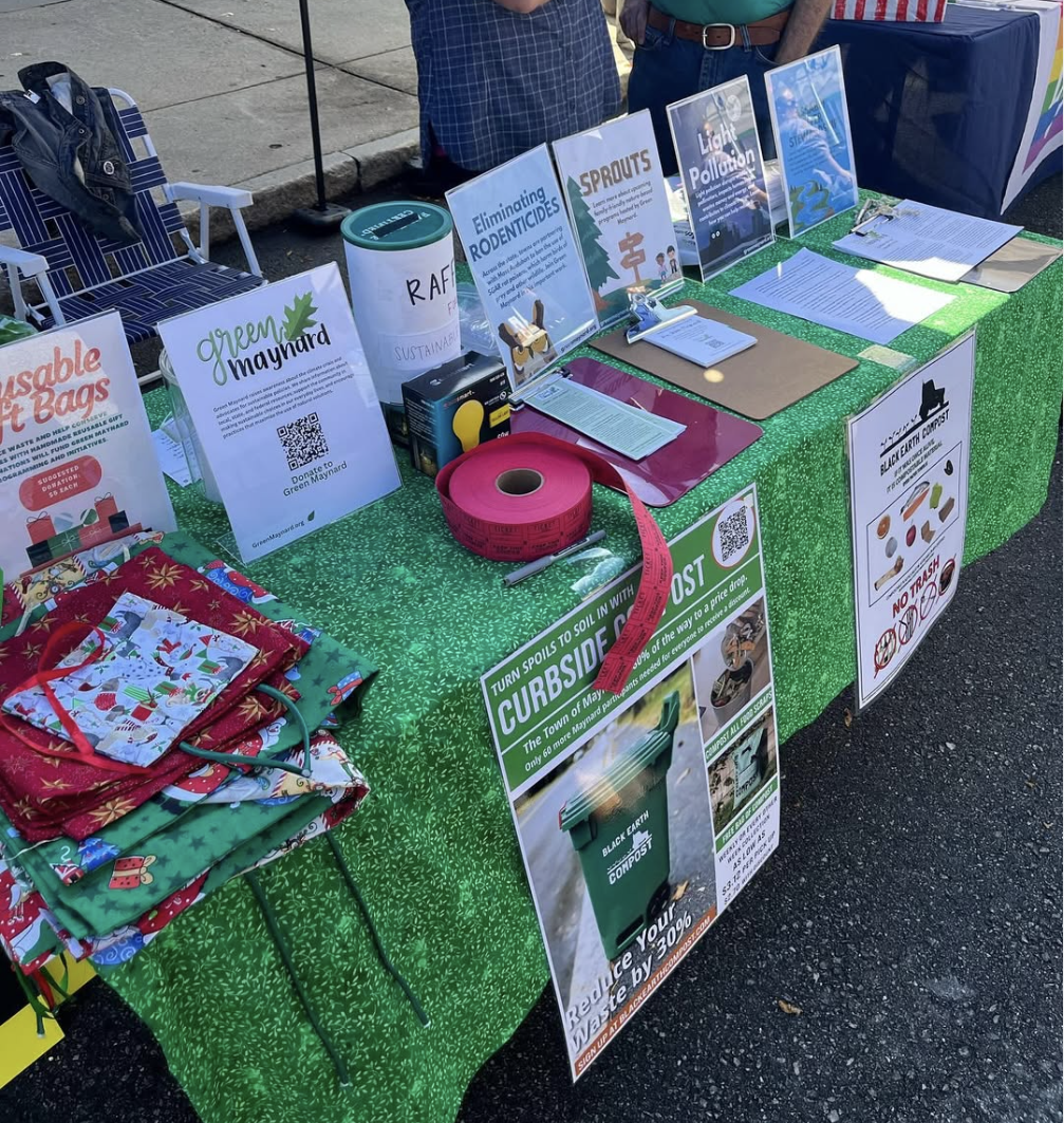 A table at an outdoor event promoting environmental sustainability through recycling and composting, with informational posters, fabric gift bags, and donation signs.