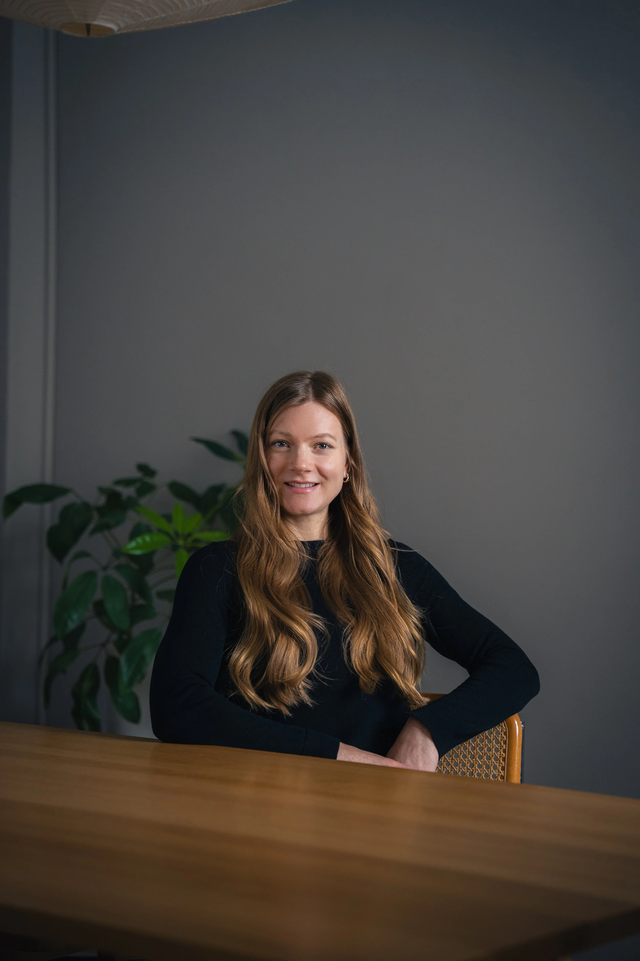 A woman with long wavy brown hair, wearing a black top, sitting at a wooden table in front of a gray wall with a green potted plant behind her, smiling at the camera.
