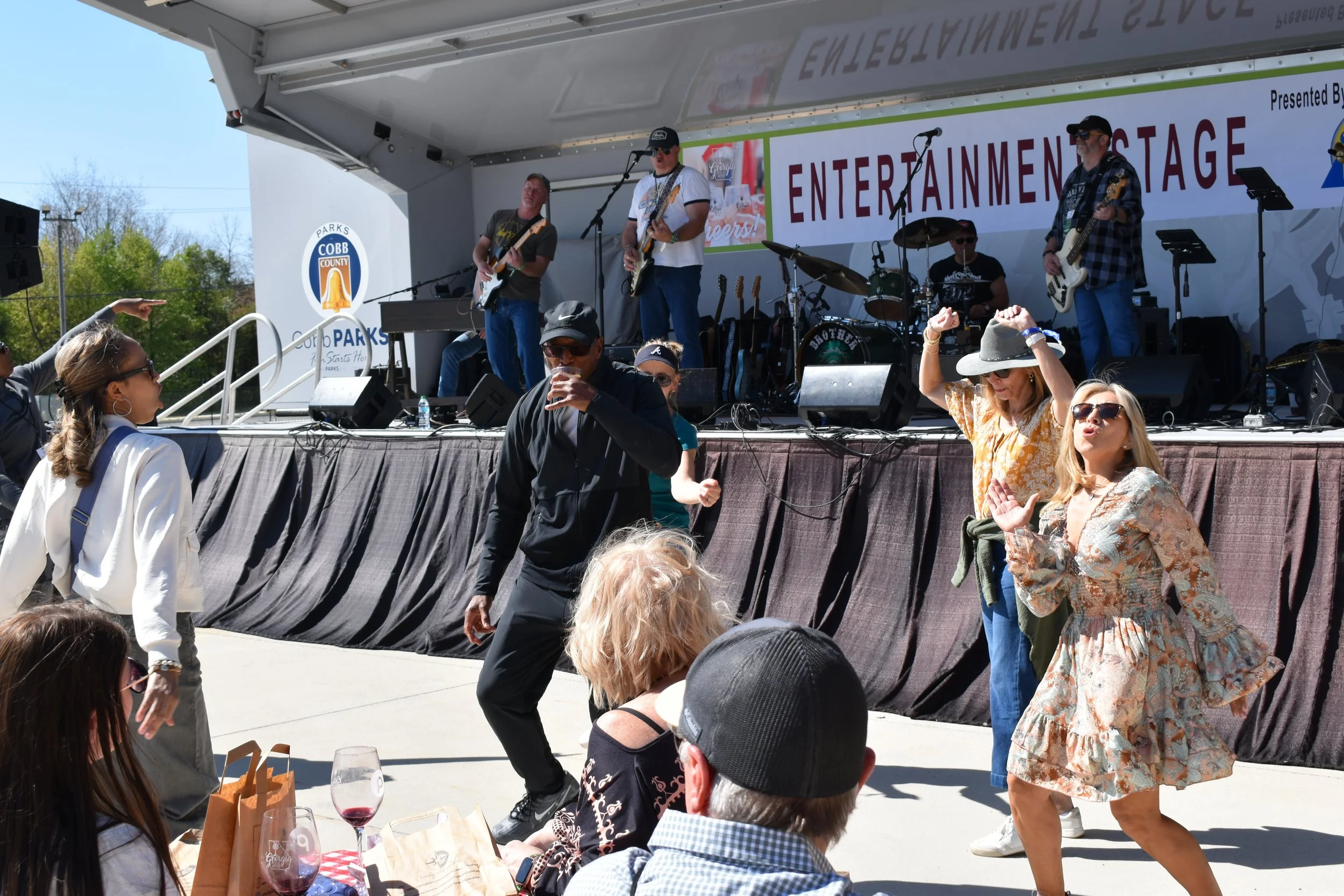 A live outdoor band performing on stage at a community event. The band has four members: three men playing guitars and one man on drums. In front of the stage, people are dancing and enjoying the music, including women in sun hats and sunglasses. Some audience members are sitting at tables with drinks.