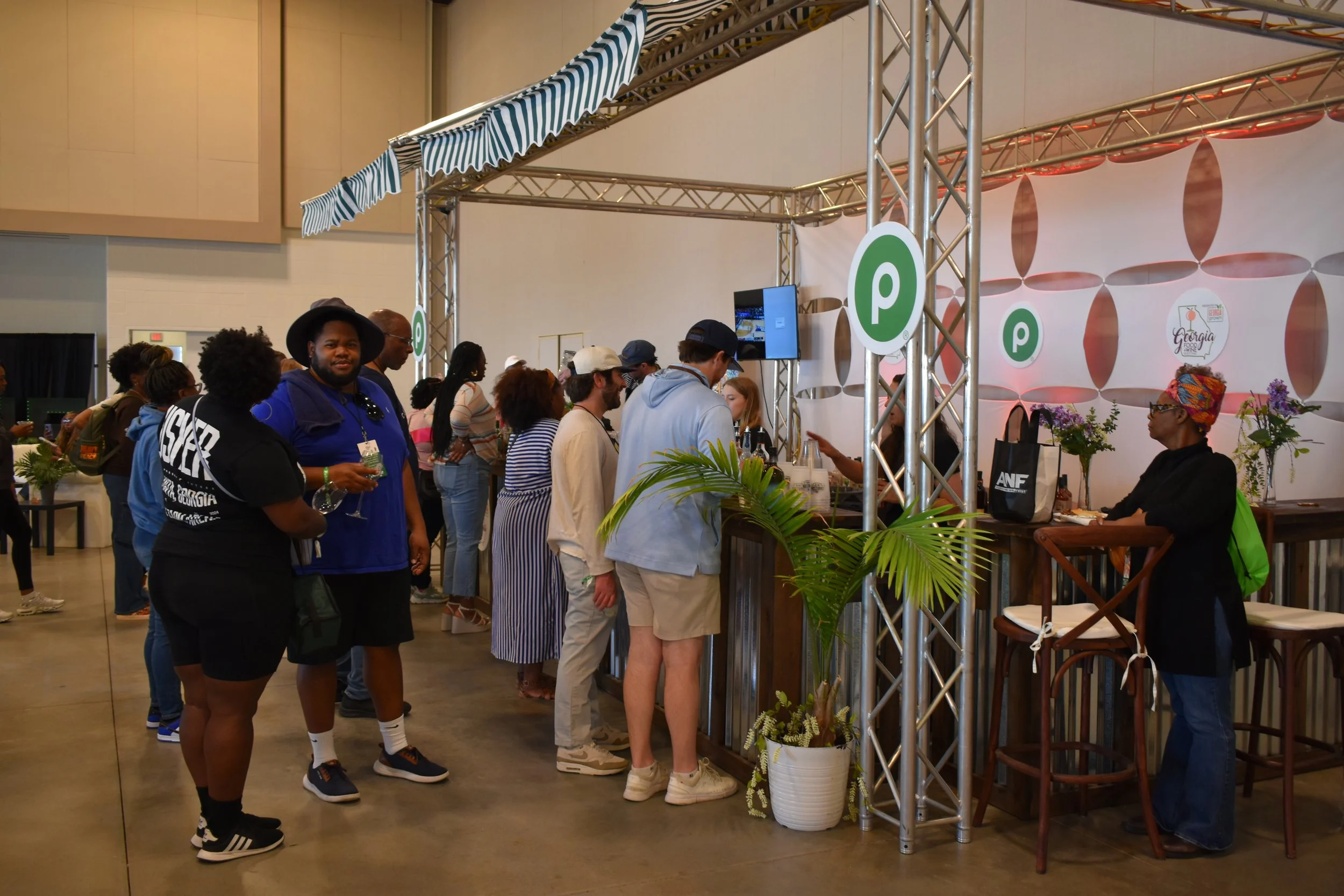People standing in line at a food or beverage stand at an indoor event, with a bar counter, decorative plants, and a partition with logo designs in the background.
