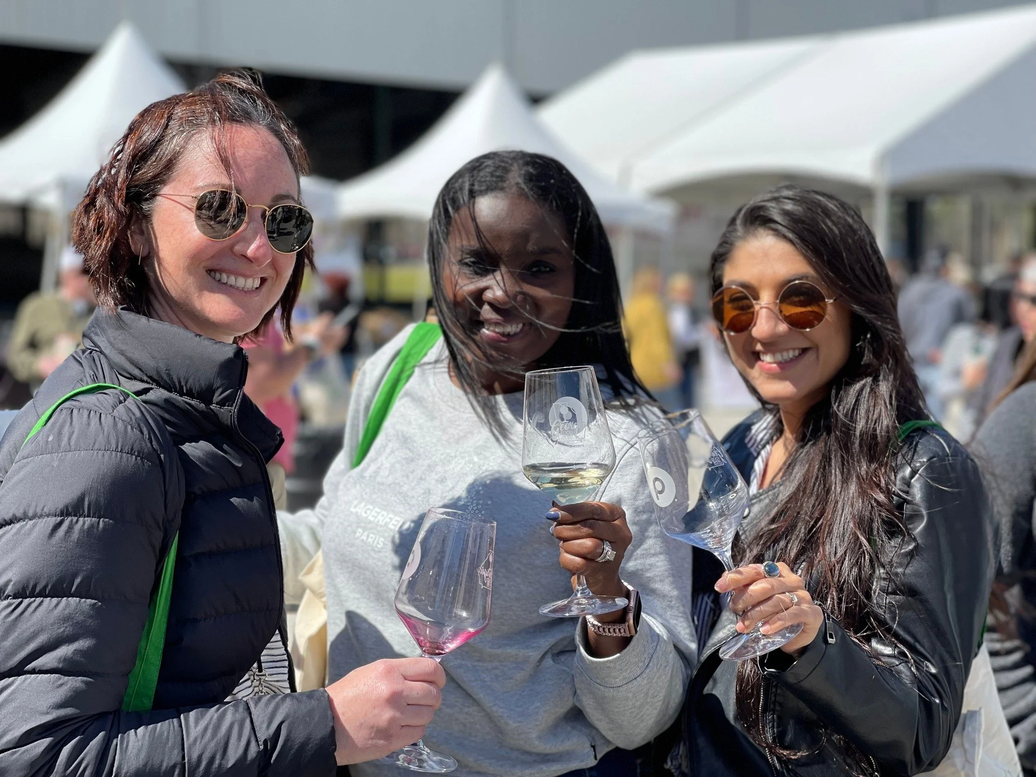 Three women smiling and holding wine glasses at an outdoor event with white tents in the background.