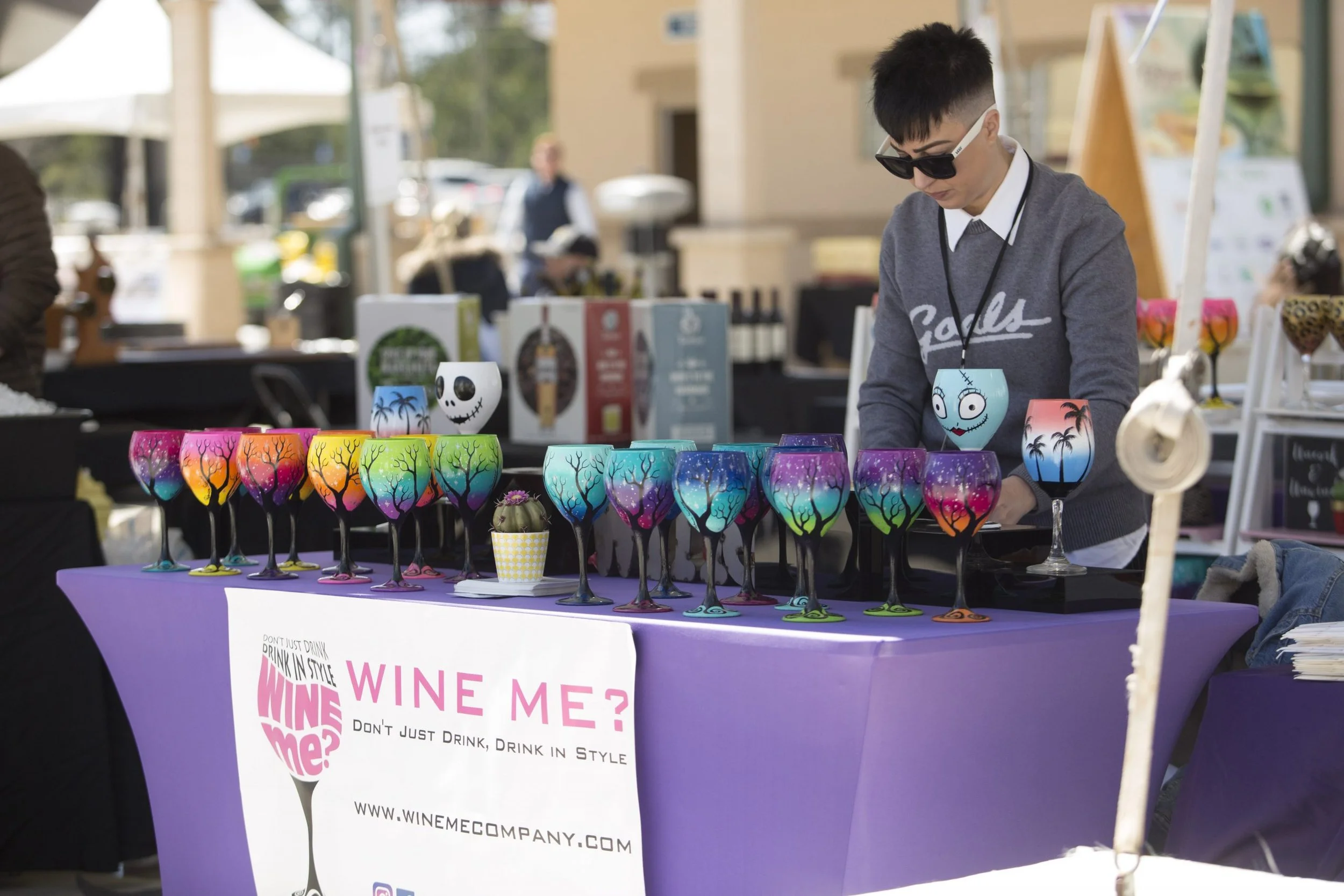 A person behind a table of colorful painted wine glasses with patterns of trees and sunsets, at an outdoor market, with a sign promoting wine in style.