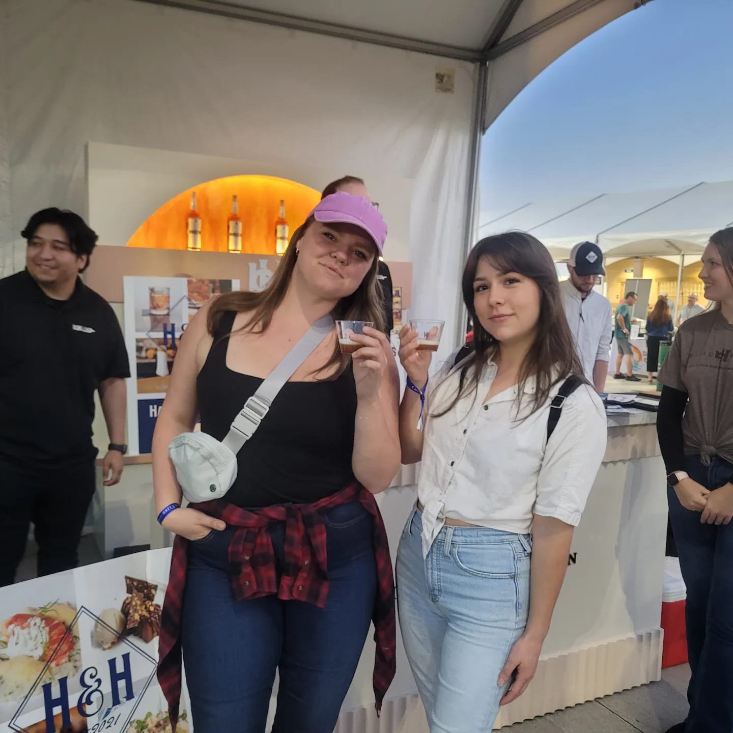 Two young women holding drinks and smiling at an outdoor event or festival, with people and a bar in the background.