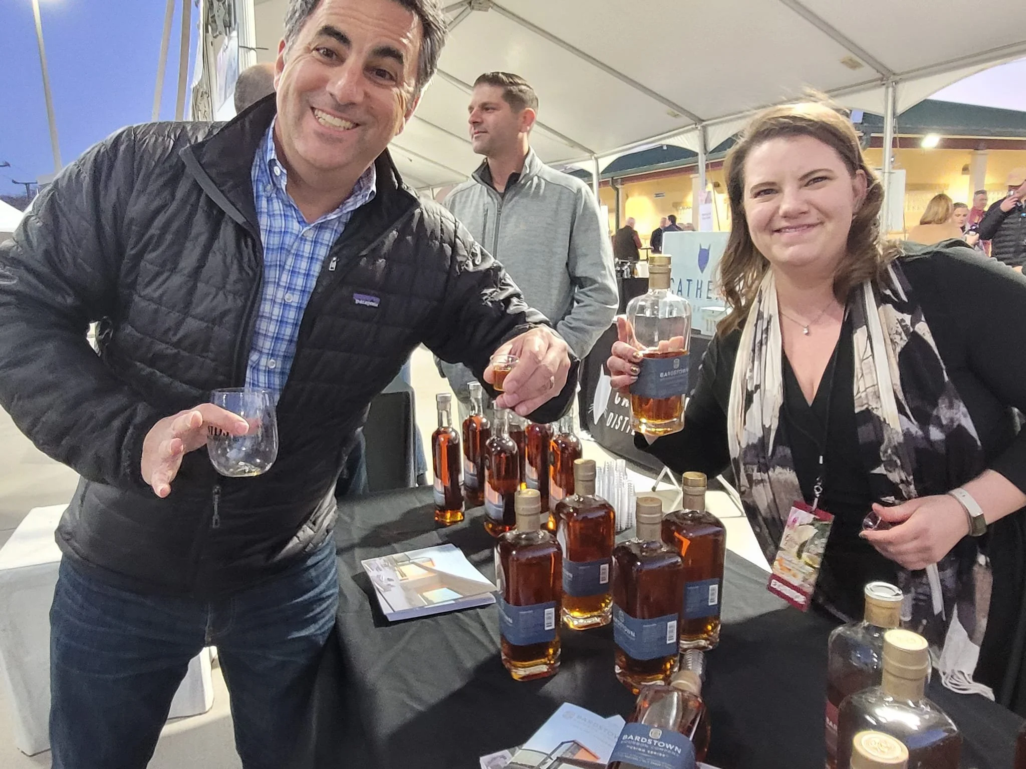 A man and woman smiling and holding drinks at a booth with bottles of whiskey at an outdoor event under a tent.