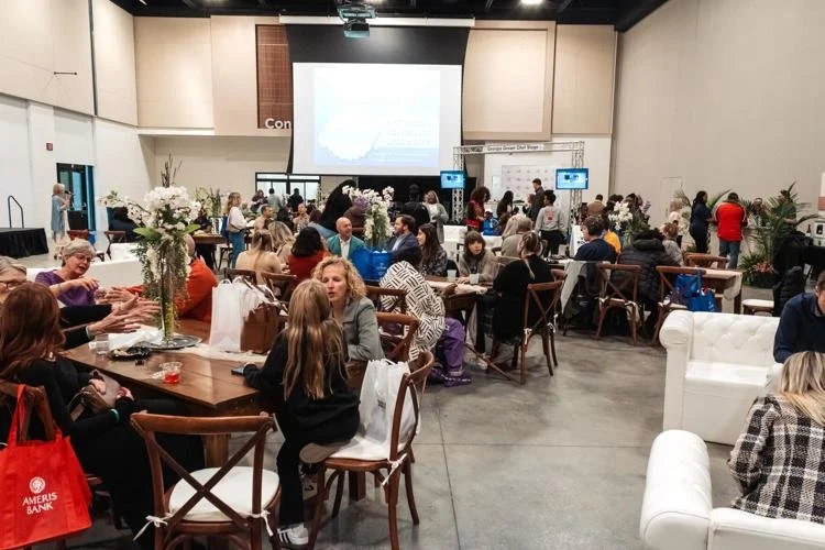 People sitting and talking at tables in a conference room with a large projection screen and multiple monitors, decorated with flower arrangements.