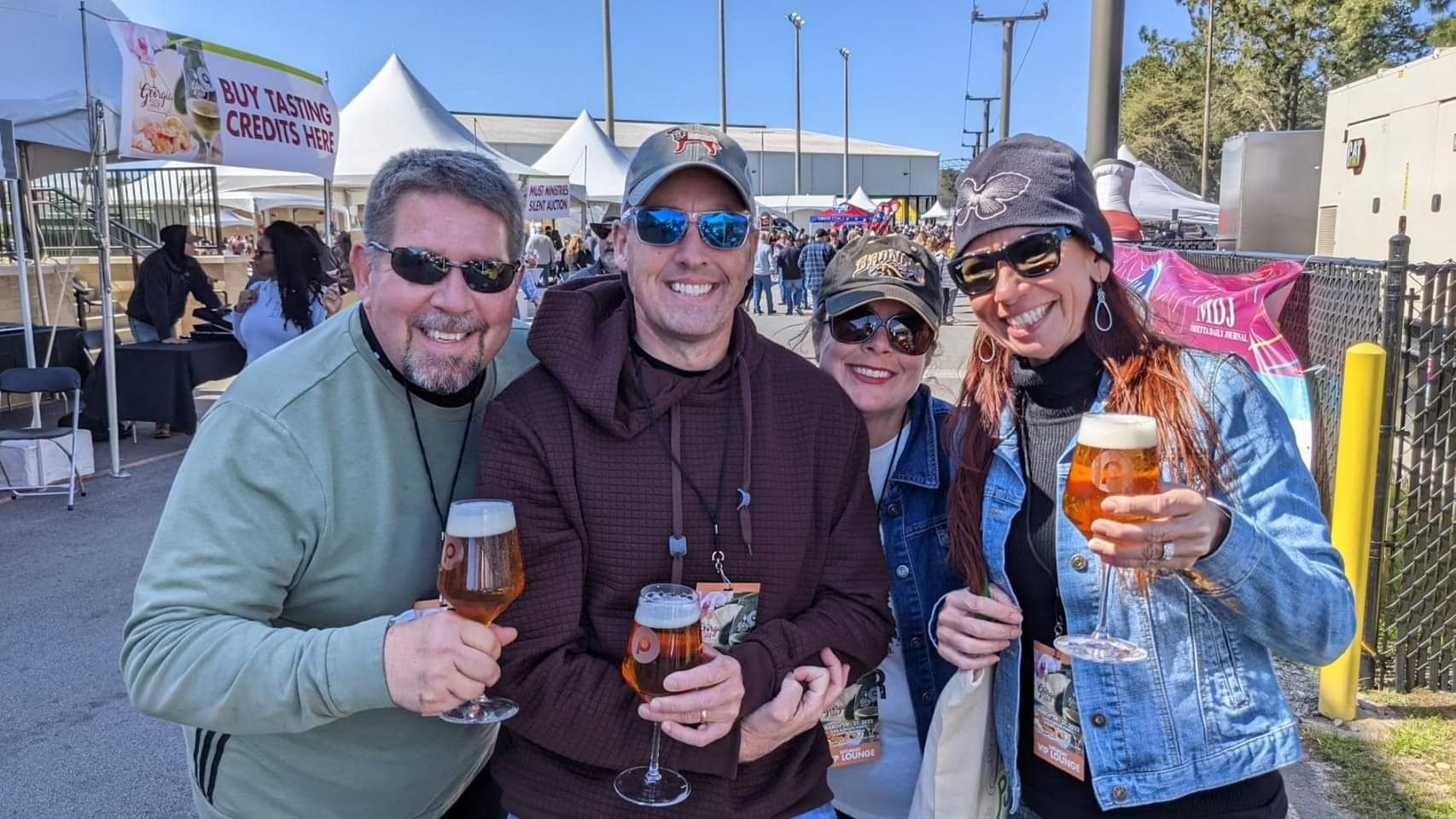 Four adults smiling and holding glasses of beer at an outdoor event with tents and people in the background.