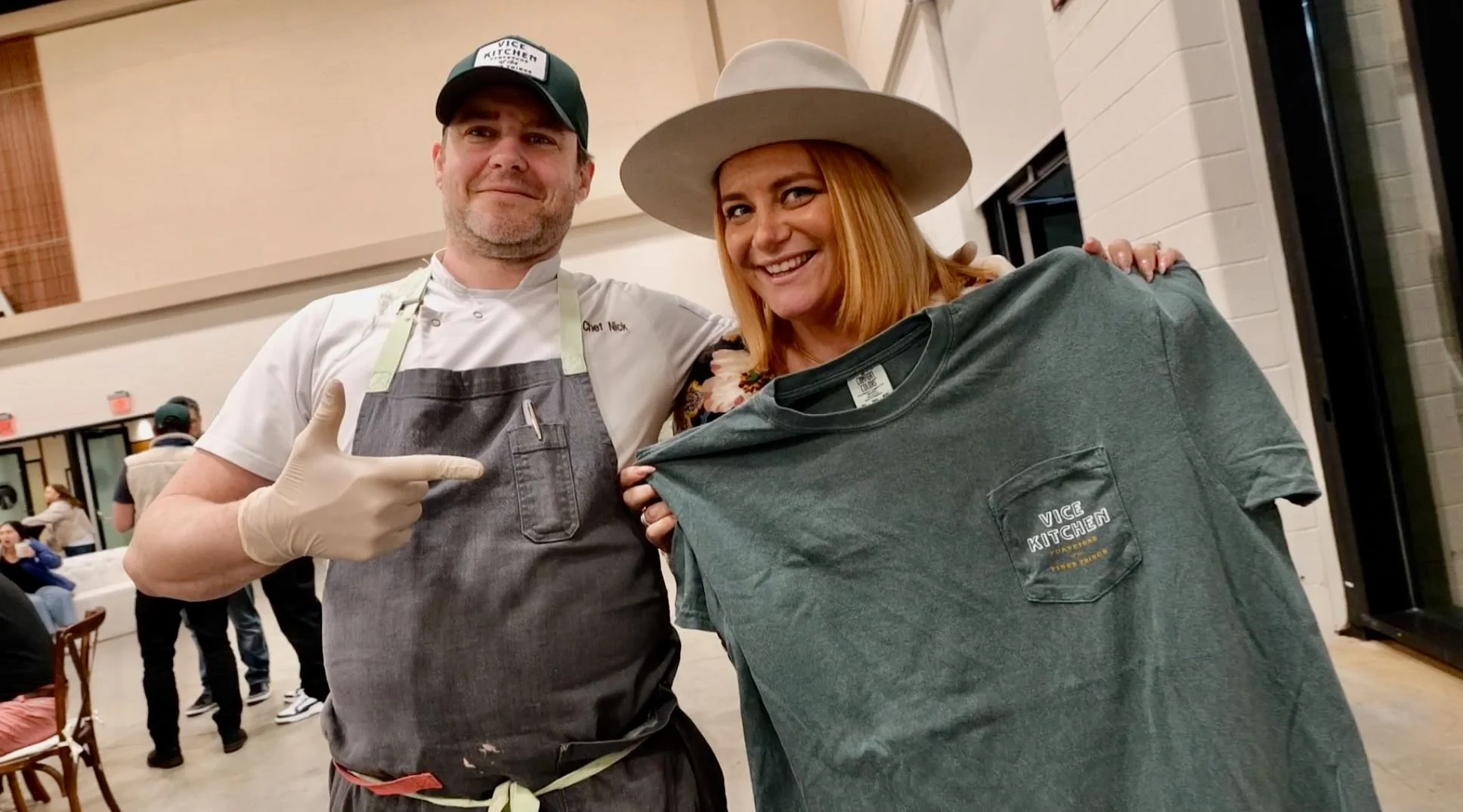 A man and woman at a social event, with the man wearing a chef's uniform, apron, and cap, and the woman holding a green T-shirt with the "VICE KITCHEN" logo. The man is pointing at the T-shirt and both are smiling.