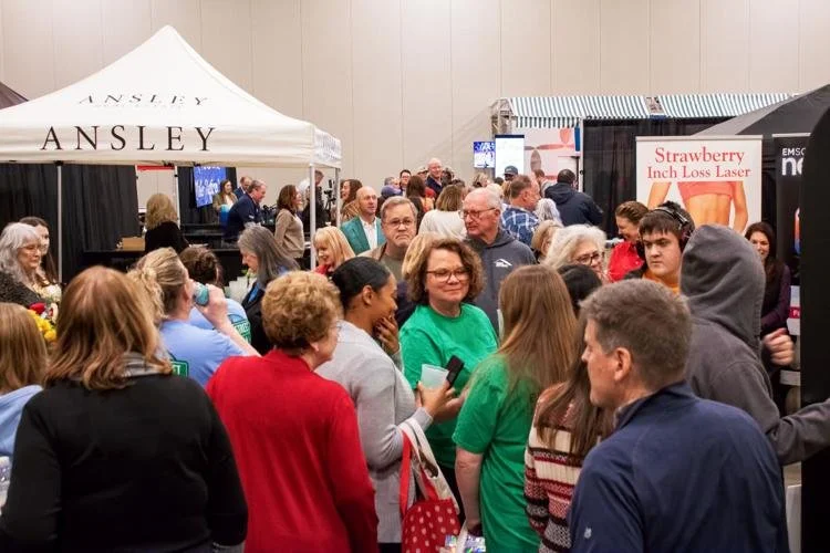 Crowd of people at an indoor event or exhibition, with tents and banners, including a strawberry laser treatment sign and a tent labeled 'Ansley'.