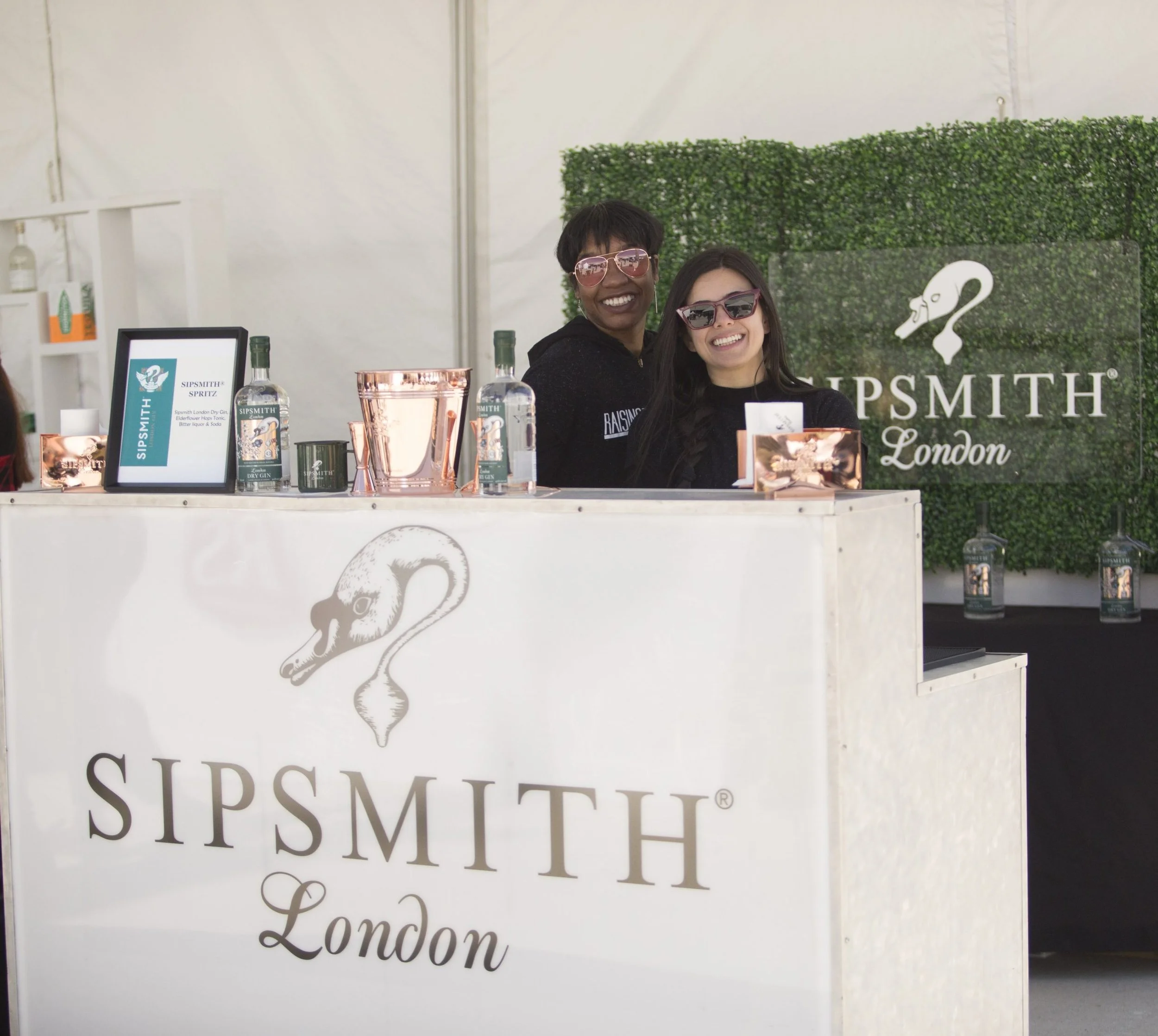 Two women smiling behind a SIPSMITH London bar counter at an event, with bottles and a framed sign on the counter and a green hedge backdrop with the SIPSMITH logo.
