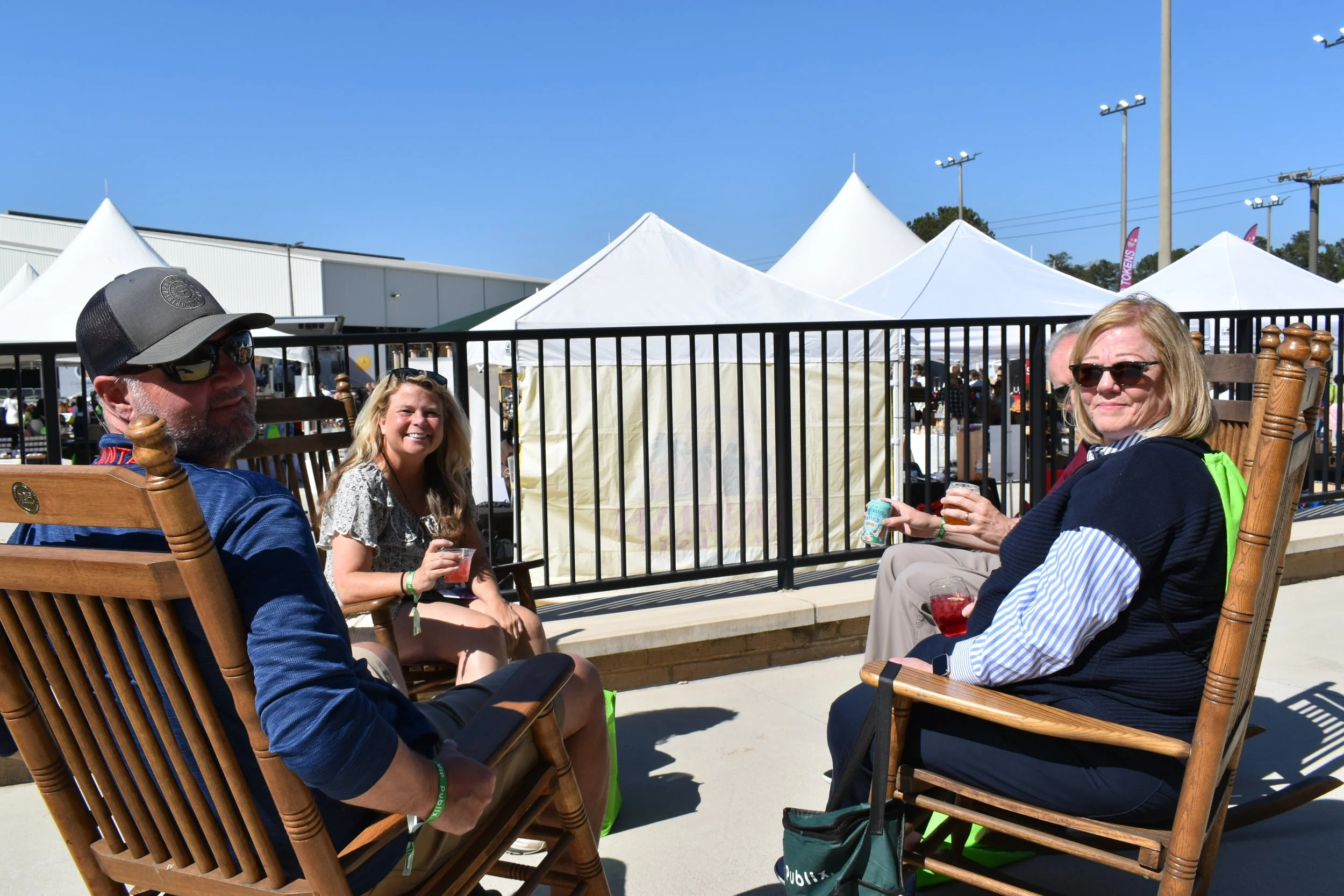 Four adults sitting on wooden rocking chairs outdoors, enjoying drinks at a festival with white tents in the background under a clear blue sky.