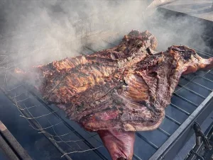A large piece of grilled meat, likely a steak or brisket, on a barbecue grill with smoke rising from it.