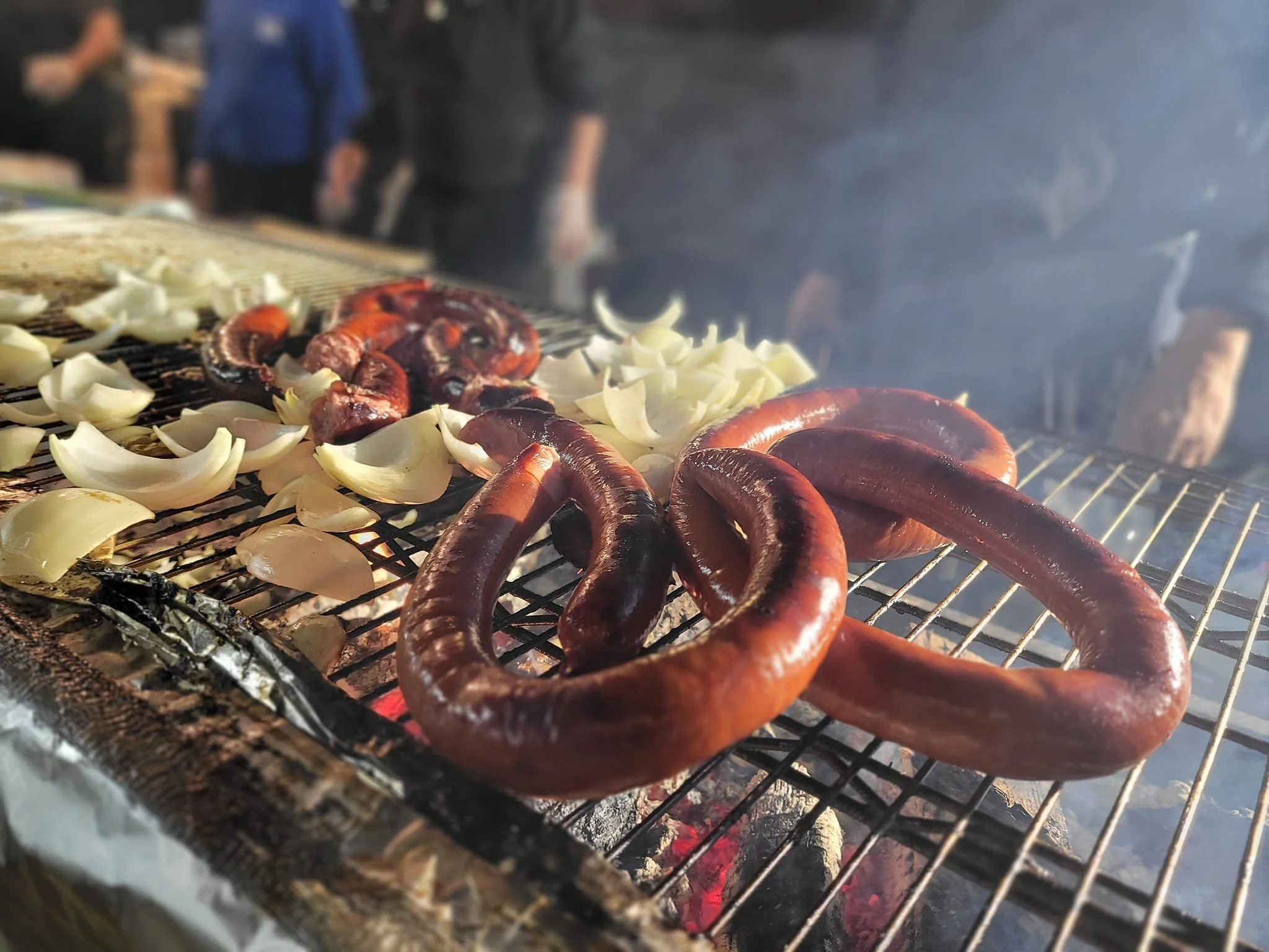 Sausages, onions, and fish grilling over an open flame on a barbecue grill with smoke in the background