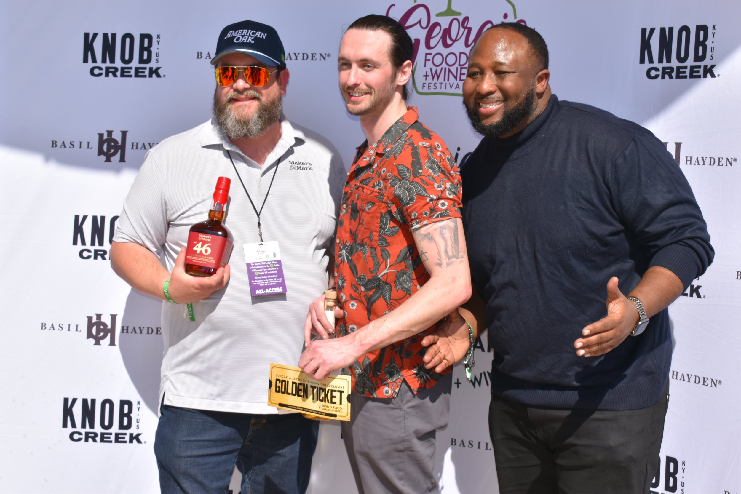 Three men standing together at an event, one holding a bottle of alcohol, with a backdrop featuring logos and text for KNOB CREEK, George Food + Wine Festival, Basil Hayden, and others.