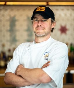A male chef wearing a white chef's coat and a black hat with yellow text, standing with arms crossed in a kitchen or restaurant setting.