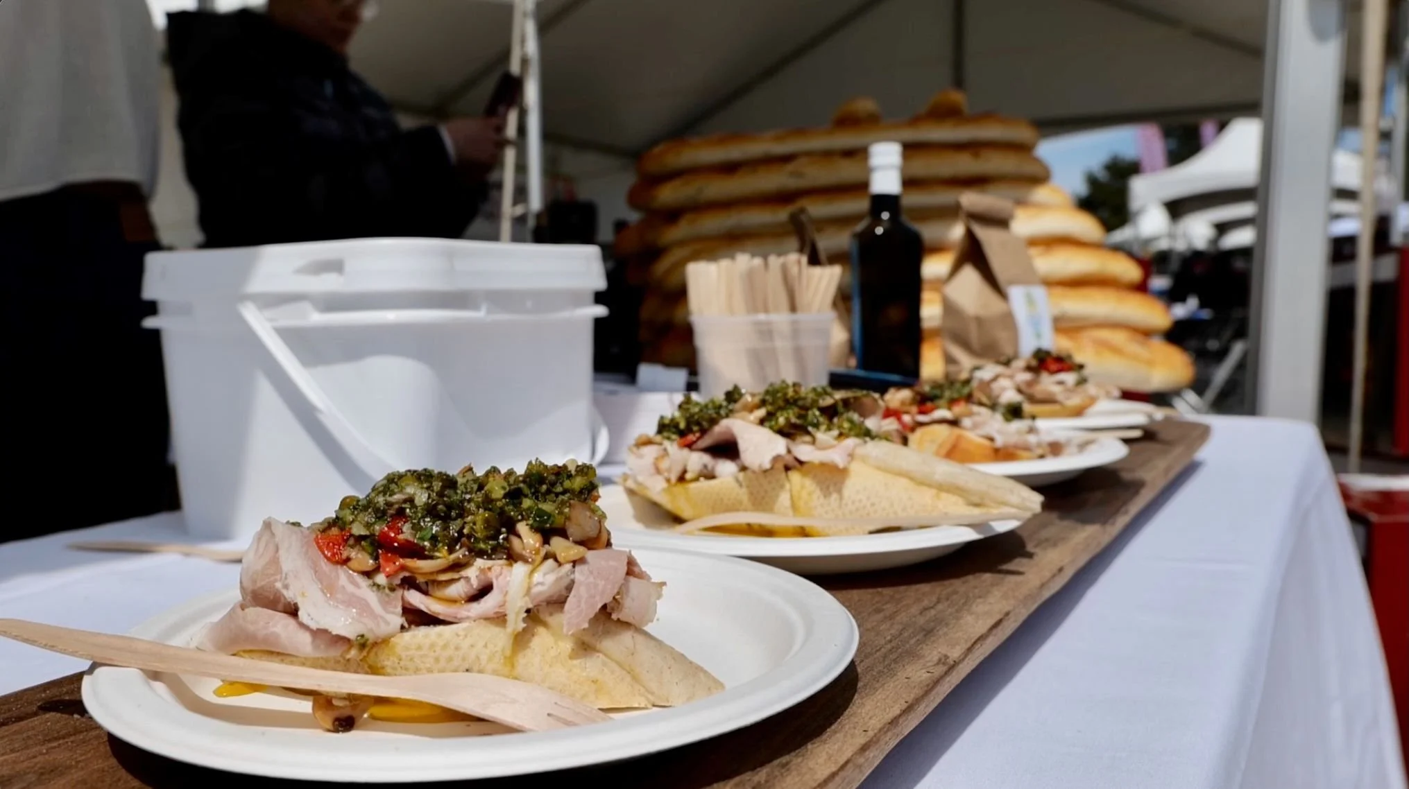 A row of plates with sandwiches featuring ham, vegetables, and bread, set on a wooden board on a table at an outdoor event. In the background are stacks of bread, a bottle, and a person.