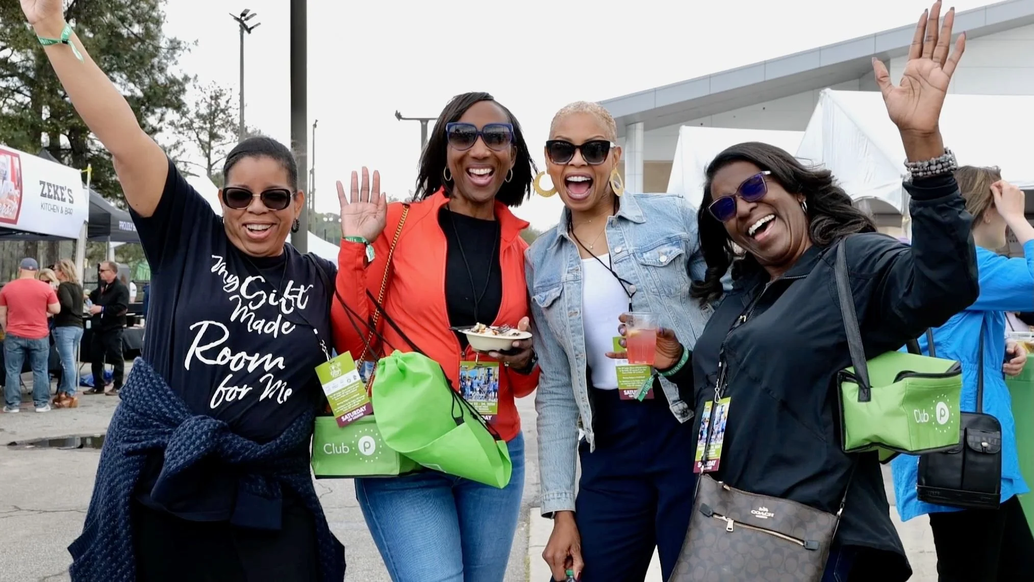 Four women at an outdoor event smiling and celebrating, waving, with booths and people in the background.