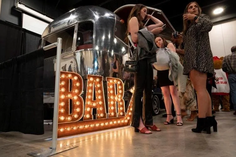 People standing near a vintage trailer with a large illuminated sign that reads 'BAR' in front of it at an indoor event.