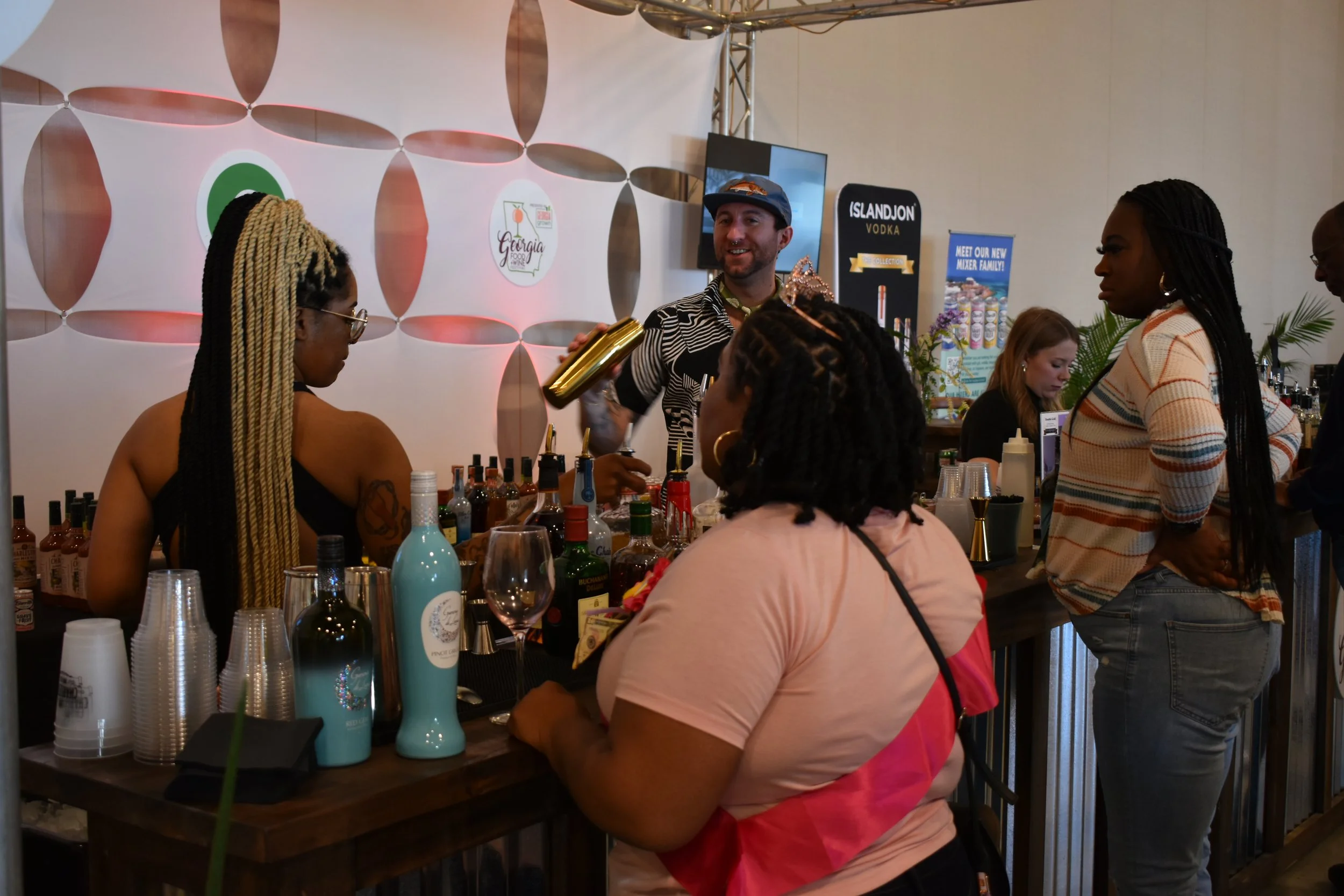 People gathered around a bar with various bottles, a bartender serving drinks, and patrons engaging with the bartender at an event or festival.