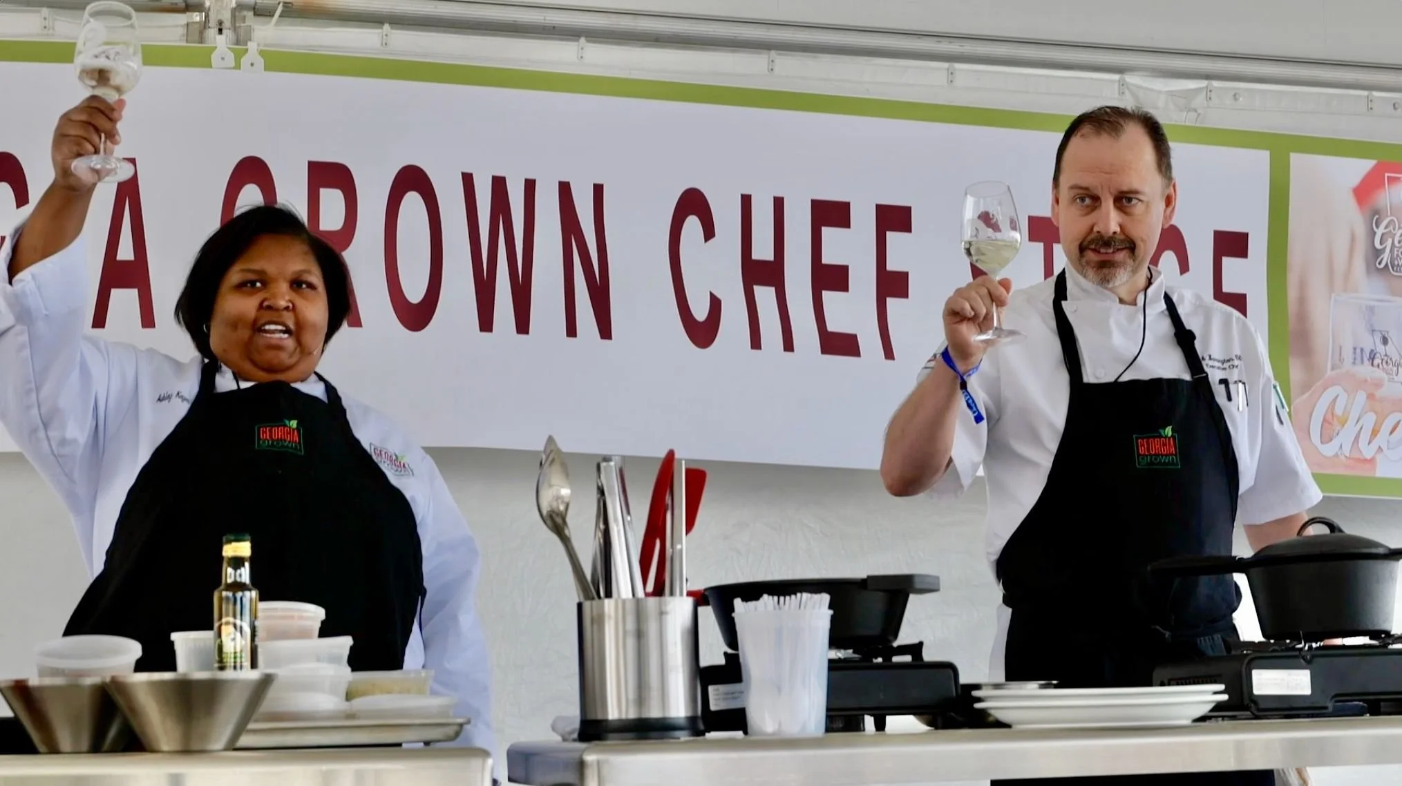 Two chefs, a woman and a man, standing at a cooking station, each holding a glass of white wine, in front of a banner that reads 'Georgia Grown Chef Stage.
