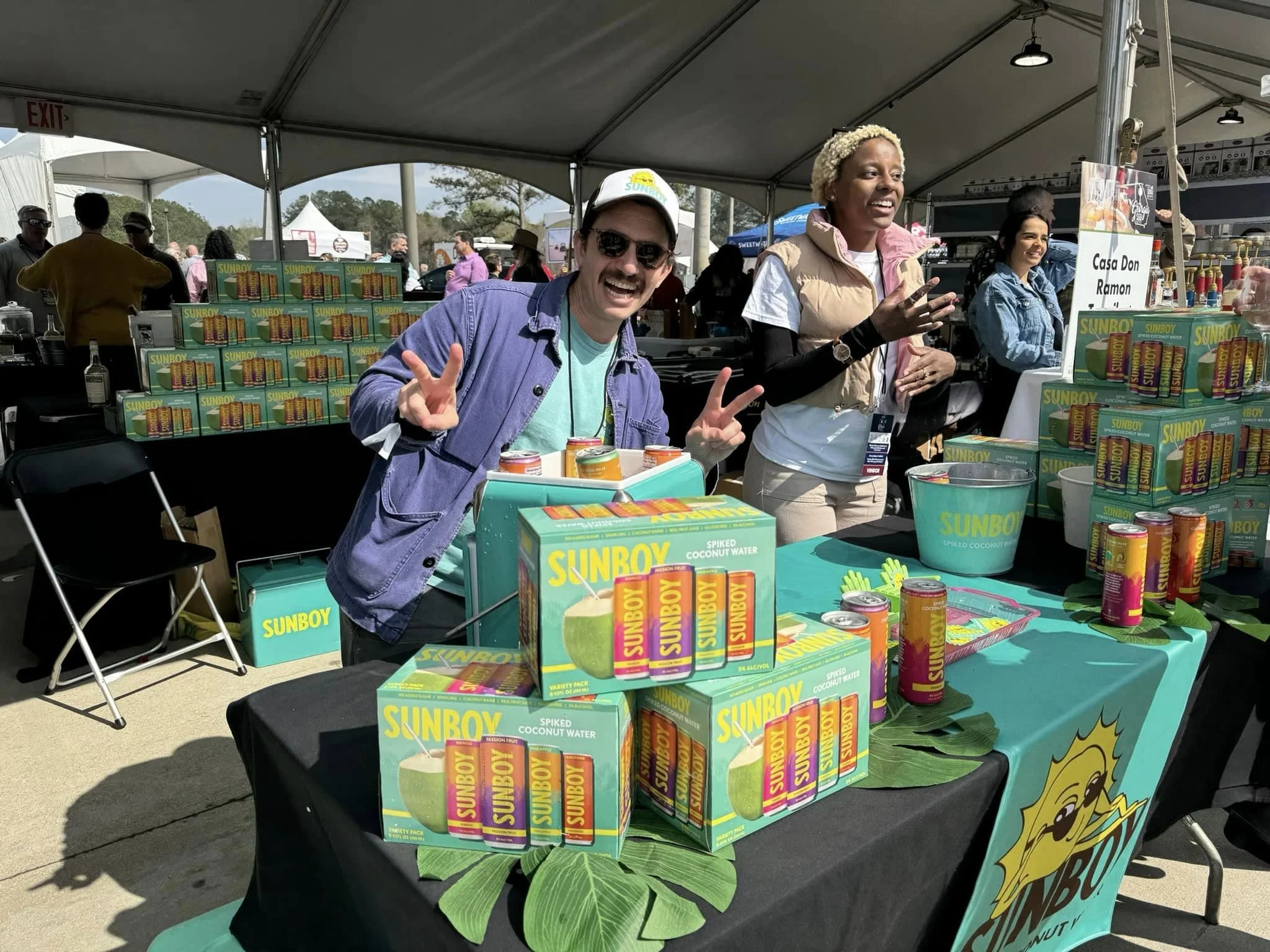 Two smiling women at a Sunboy coconut water booth at an outdoor event, with multiple boxes of Sunboy drinks on display, a table covered with a Sunboy branded cloth, and other booths and people in the background.