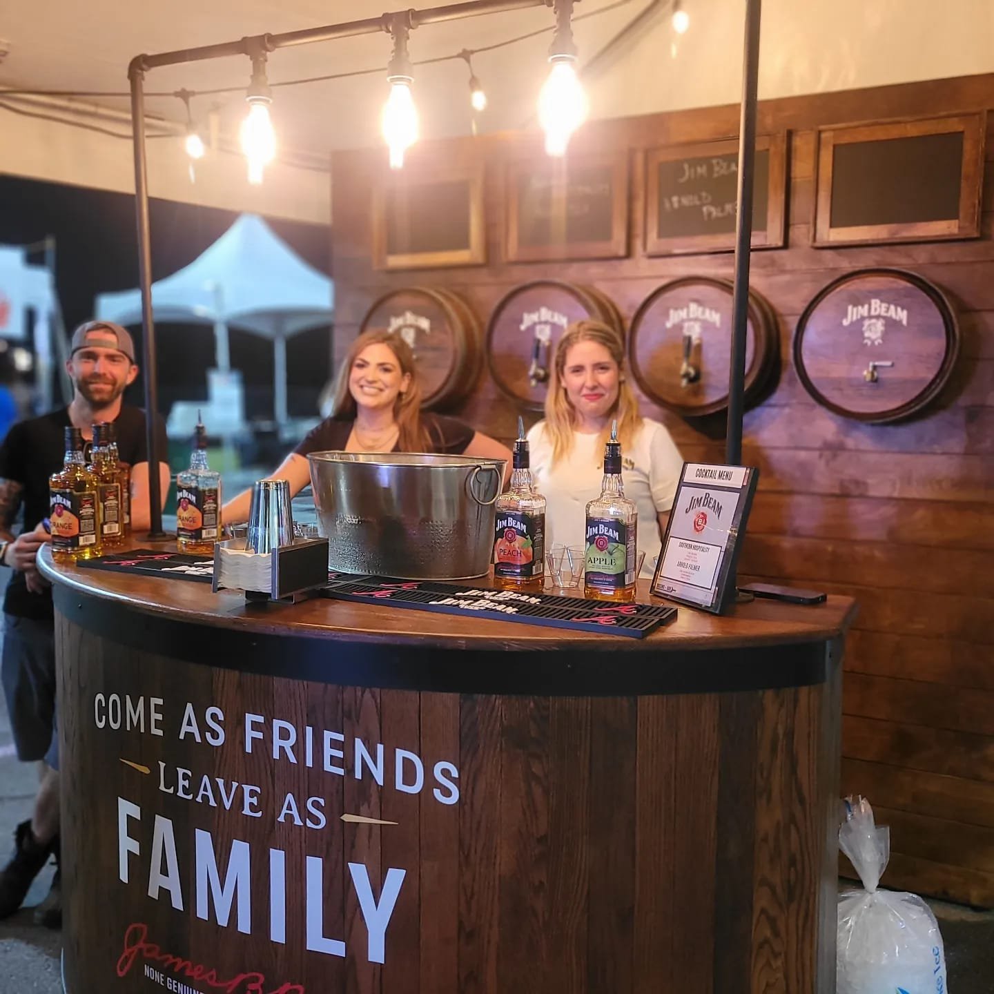 Three smiling people behind a Jim Beam branded bar at an outdoor event. The bar has a sign reading "Come as friends, leave as family". Bottles of Jim Beam whiskey and cocktail menus are on the counter, with warm hanging lights overhead and wooden barrels in the background.