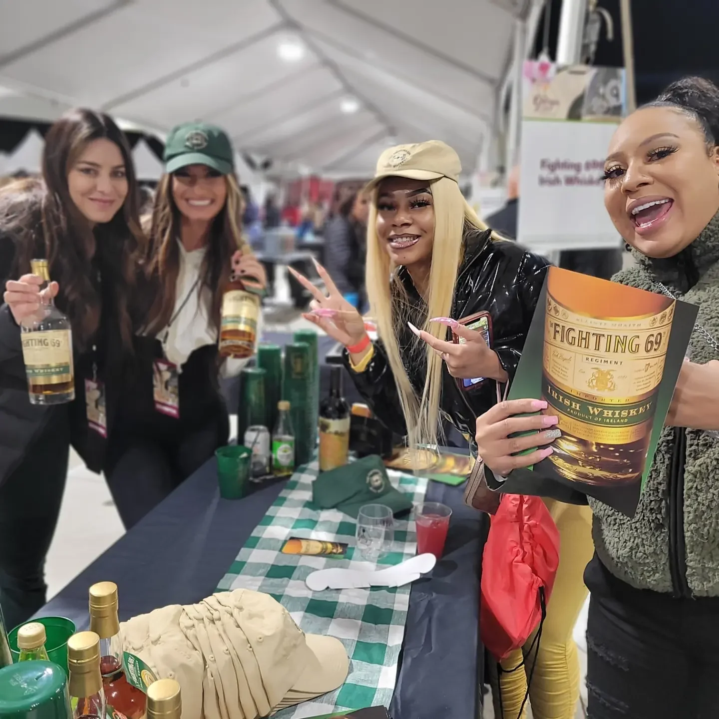 Four women at a booth celebrating with bottles of whiskey, glasses, and a cake shaped like a hat, under a white tent at an event.