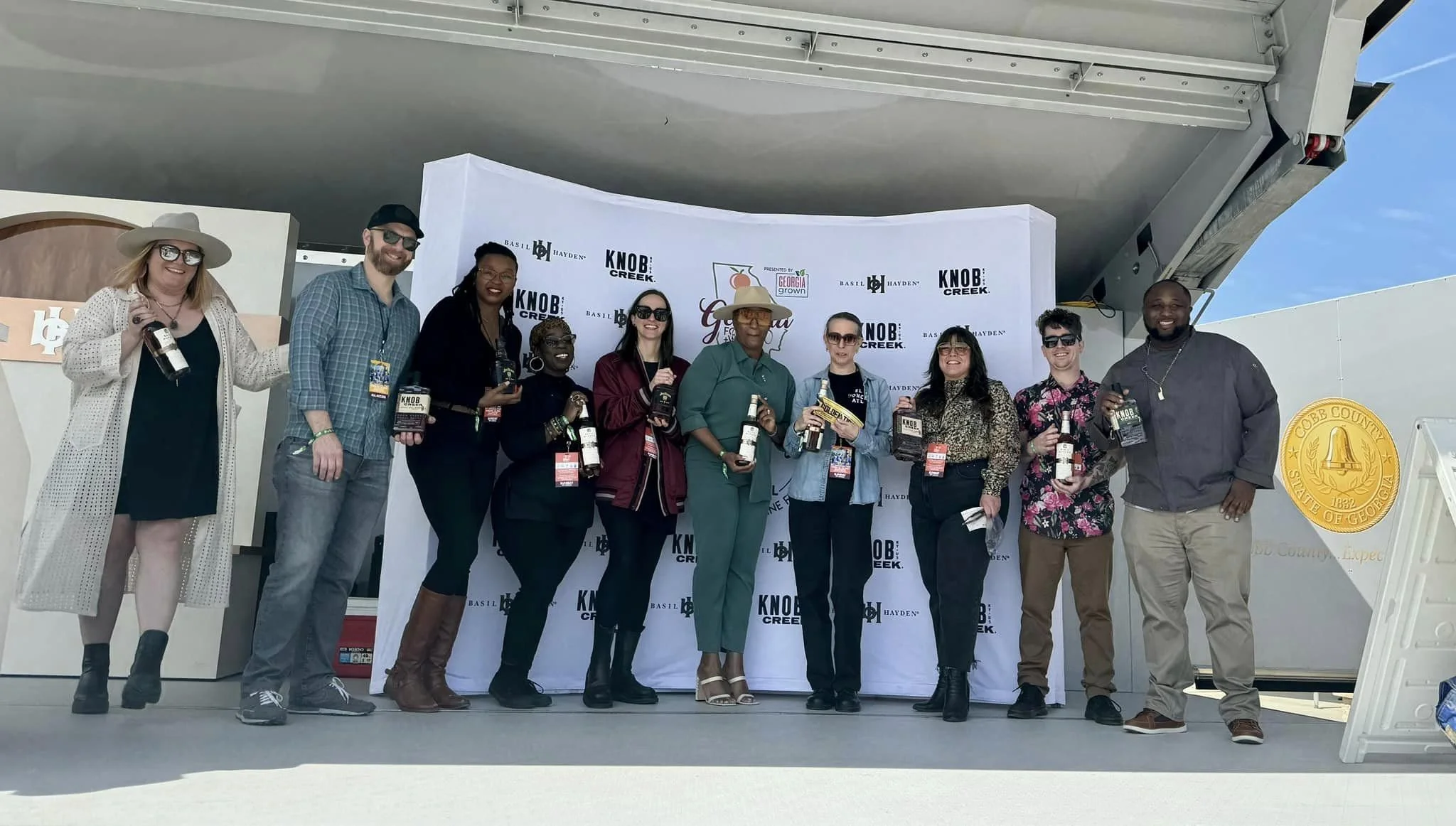 Group of ten diverse people standing on a stage holding bottles of alcohol, with a white backdrop featuring logos and text, outdoors under a clear blue sky.