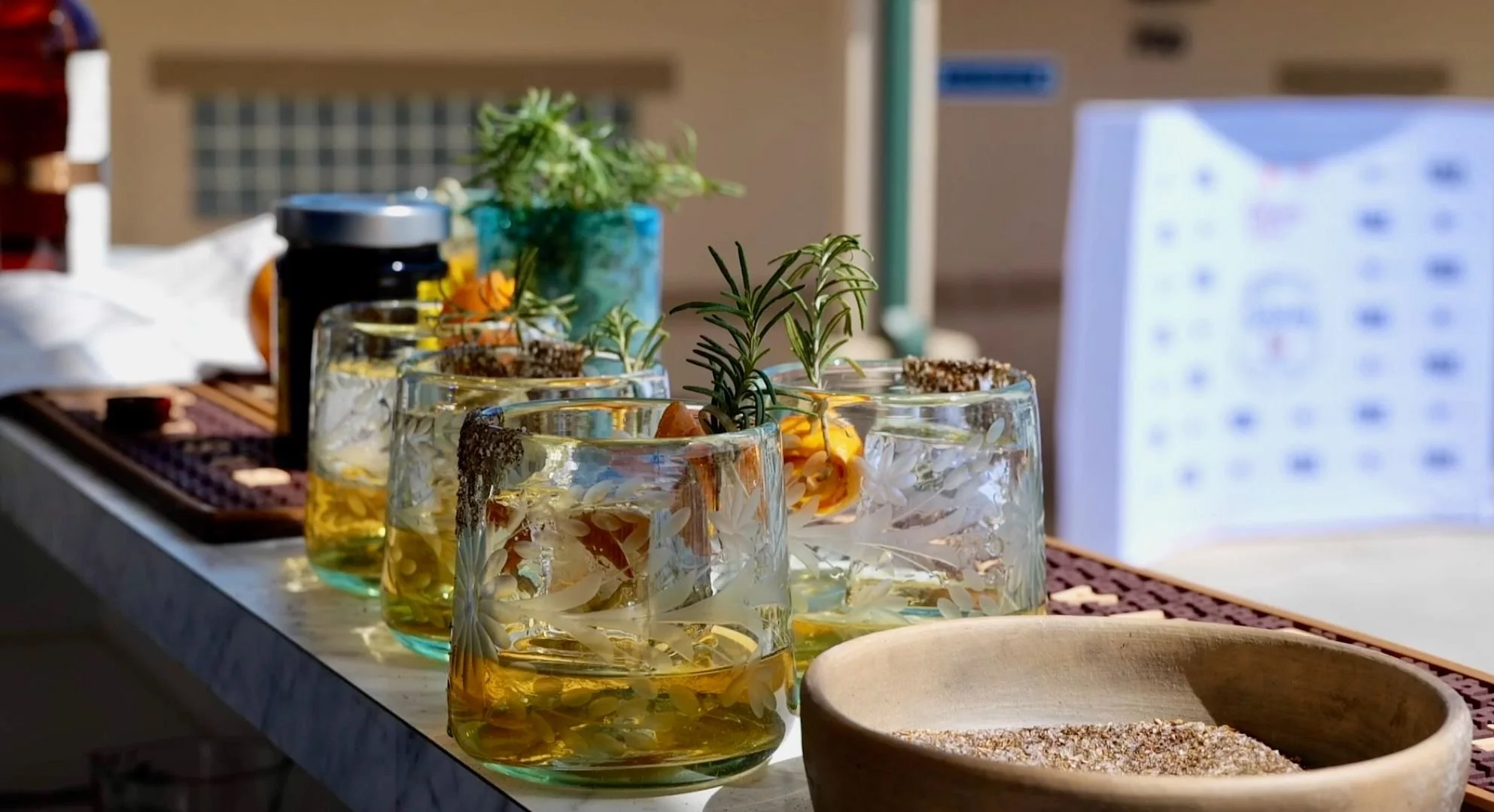 Six glass cups filled with a yellow beverage, decorated with various herbs and flowers, placed on a marble surface outdoors, with a wooden bowl containing a grain or powder, and potted plants in the background.