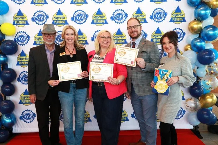 Group of five people standing in front of a backdrop with logos, holding certificates and a book, with balloons on the sides, celebrating an award event.