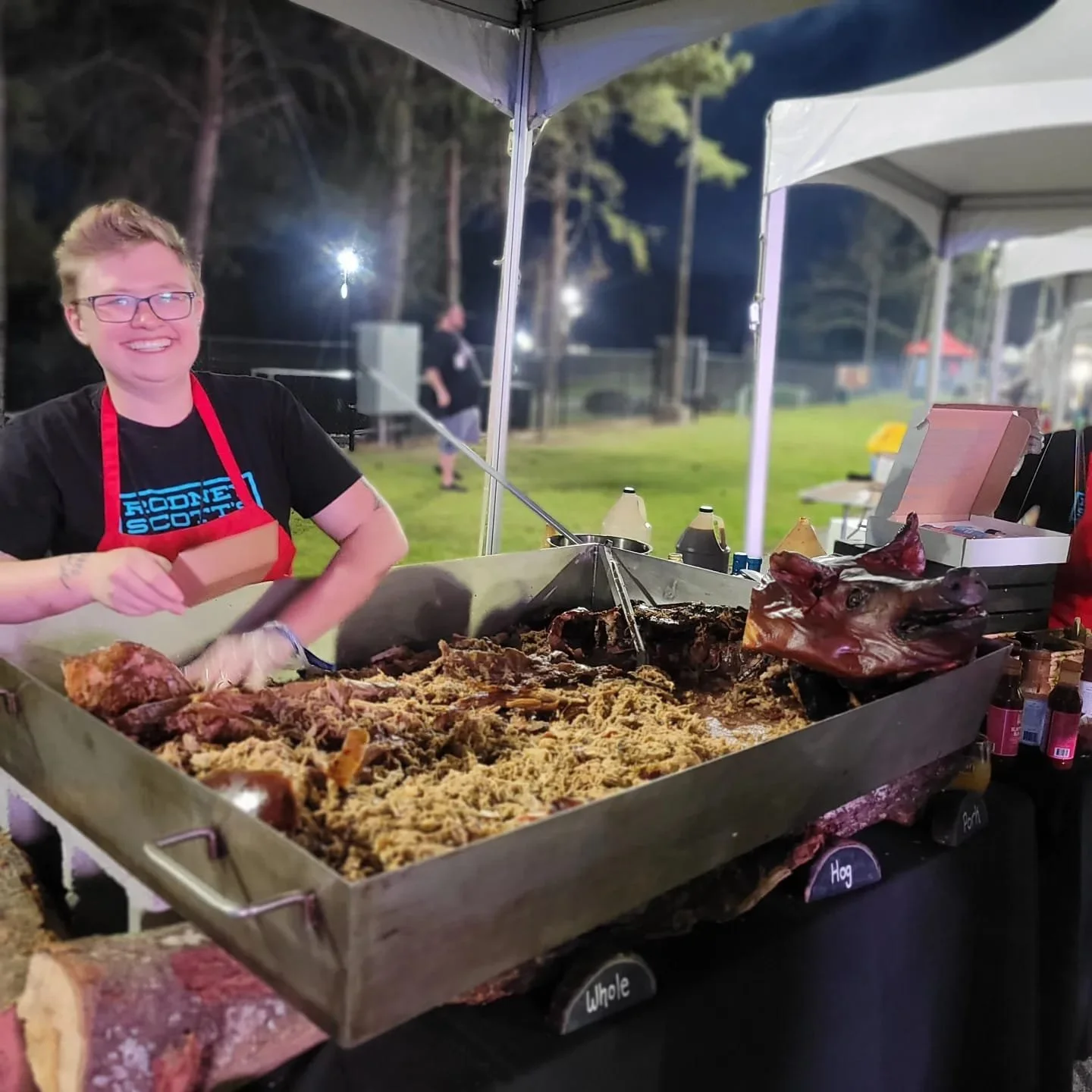 A smiling woman wearing glasses, a black t-shirt, and a red apron standing at a food stall with a large tray of cooked meat and shredded pork, labeled with small signs. The background shows a park or outdoor event at night.