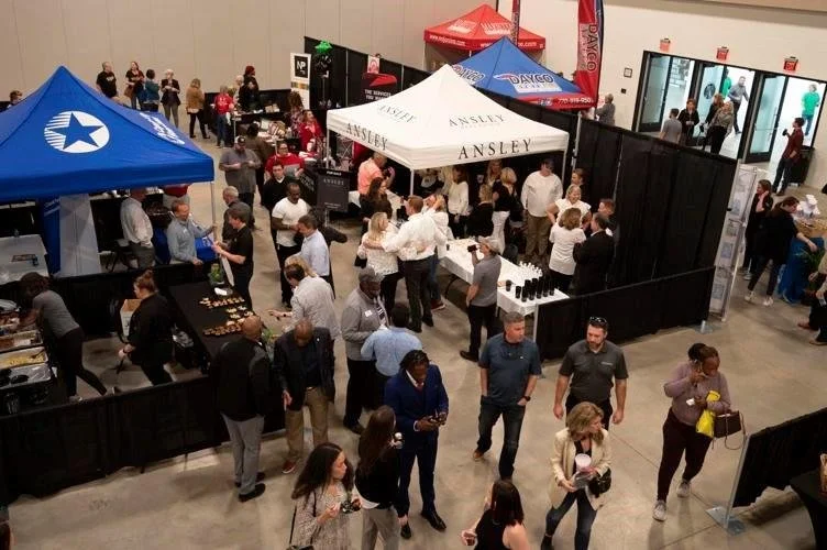 People attending an indoor trade show or convention, with multiple booths and tents. Some booths display branding such as 'Ansley,' 'Dango,' and a tent with a blue star logo. Attendees are walking around, exploring displays, and engaging with exhibitors.