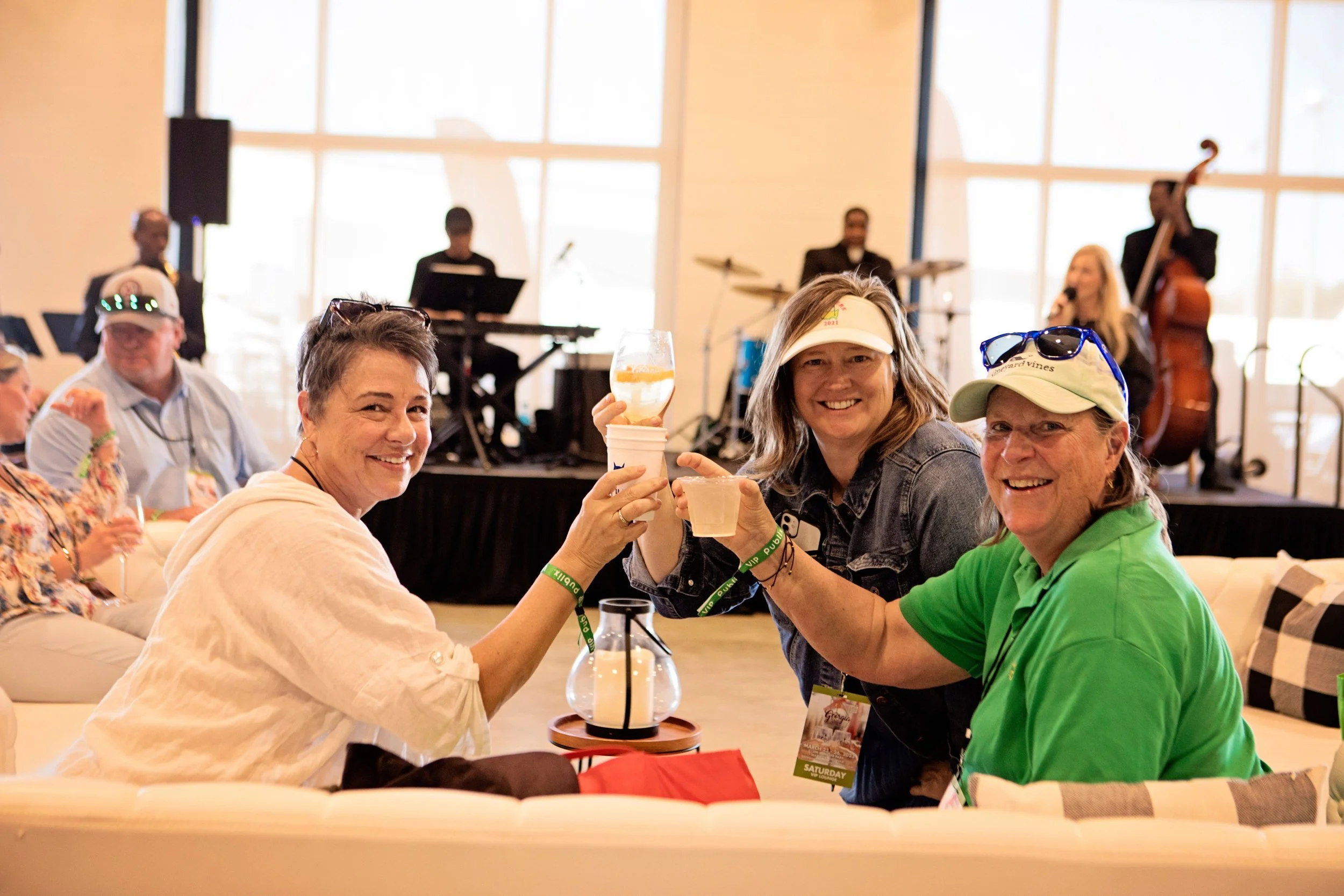 Three women sitting on a beige couch at a social event, toasting with drinks, with a band performing on stage in the background.
