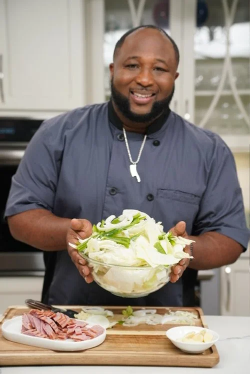A man smiling and holding a large bowl of sliced cabbage in a kitchen with a cutting board, sliced ham, and a small bowl of butter on the counter.