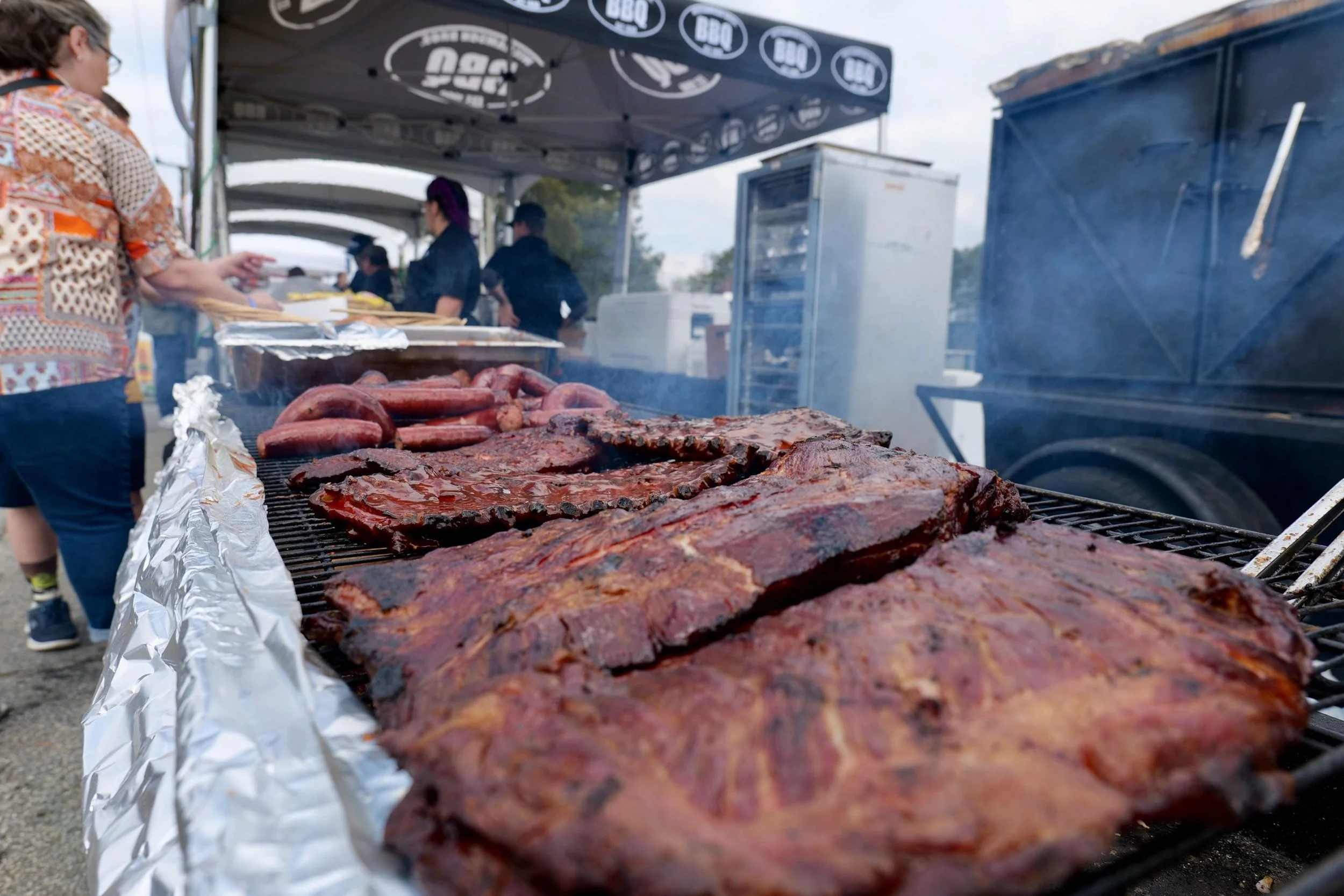 Barbecue food being grilled at an outdoor event, including ribs and sausages, with people in the background under a canopy.